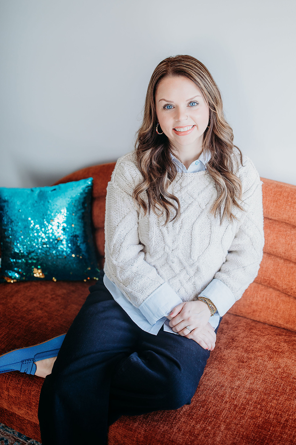 A woman with long wavy brown hair, blue eyes, and light skin sitting on an orange sofa with a sparkly teal pillow behind her, wearing a cream cable knit sweater, white shirt, and dark pants, smiling at the camera.