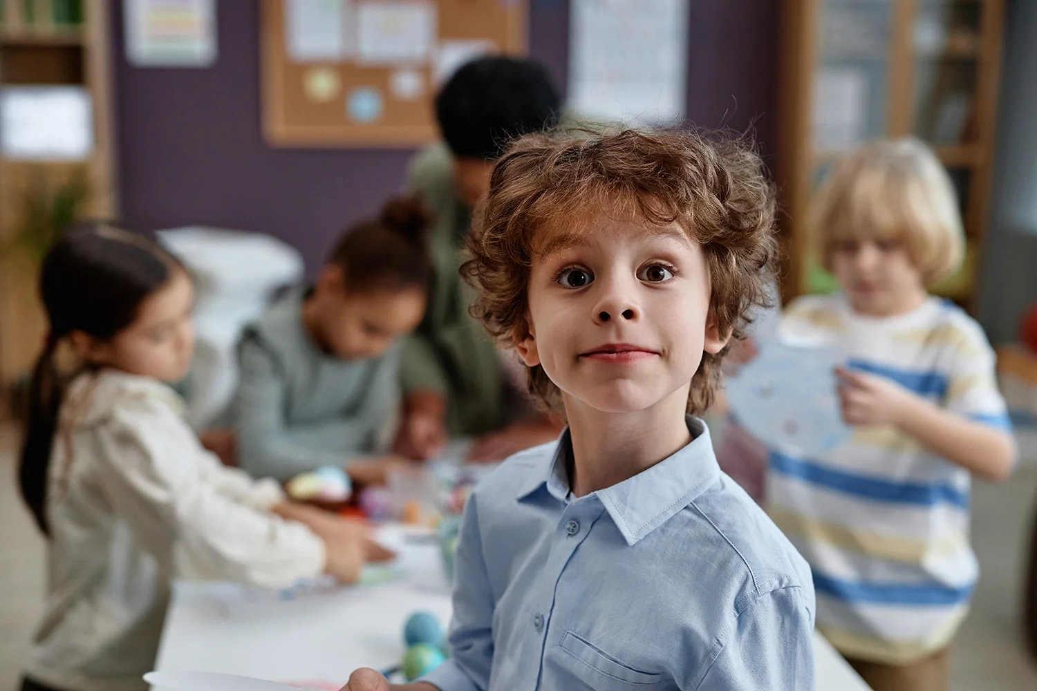 A young boy with curly hair and a light blue shirt looking at the camera in a classroom with other children in the background.