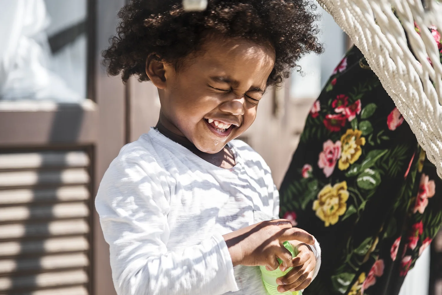 A young boy with curly hair is smiling and laughing while holding a green squeegee, opening a bottle of bubbles, outdoors on a bright day with sunlight.