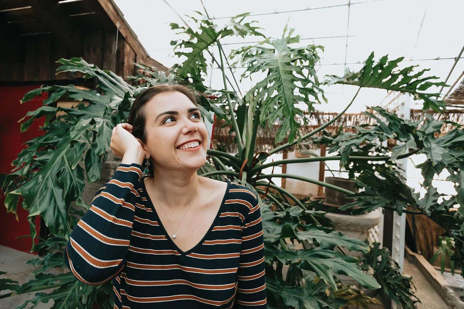 A woman smiling and looking upward, standing in front of large leafy plants inside a greenhouse.