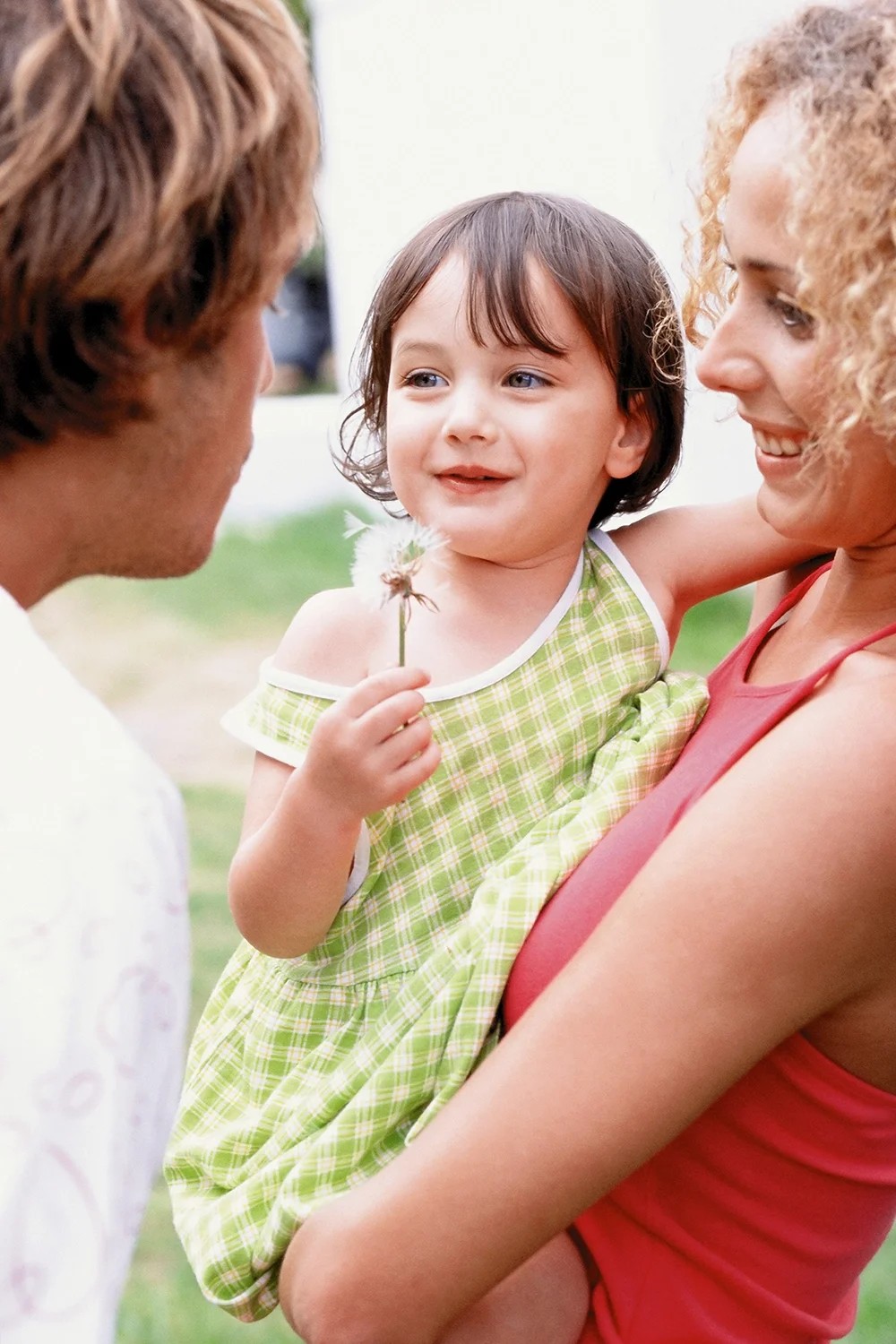 A young girl with short brown hair, wearing a green and white checkered dress, is being held by a woman with curly blonde hair dressed in a red top. The girl is holding a dandelion and is smiling at a man with brown hair who is facing her. They are outdoors with a blurred background.