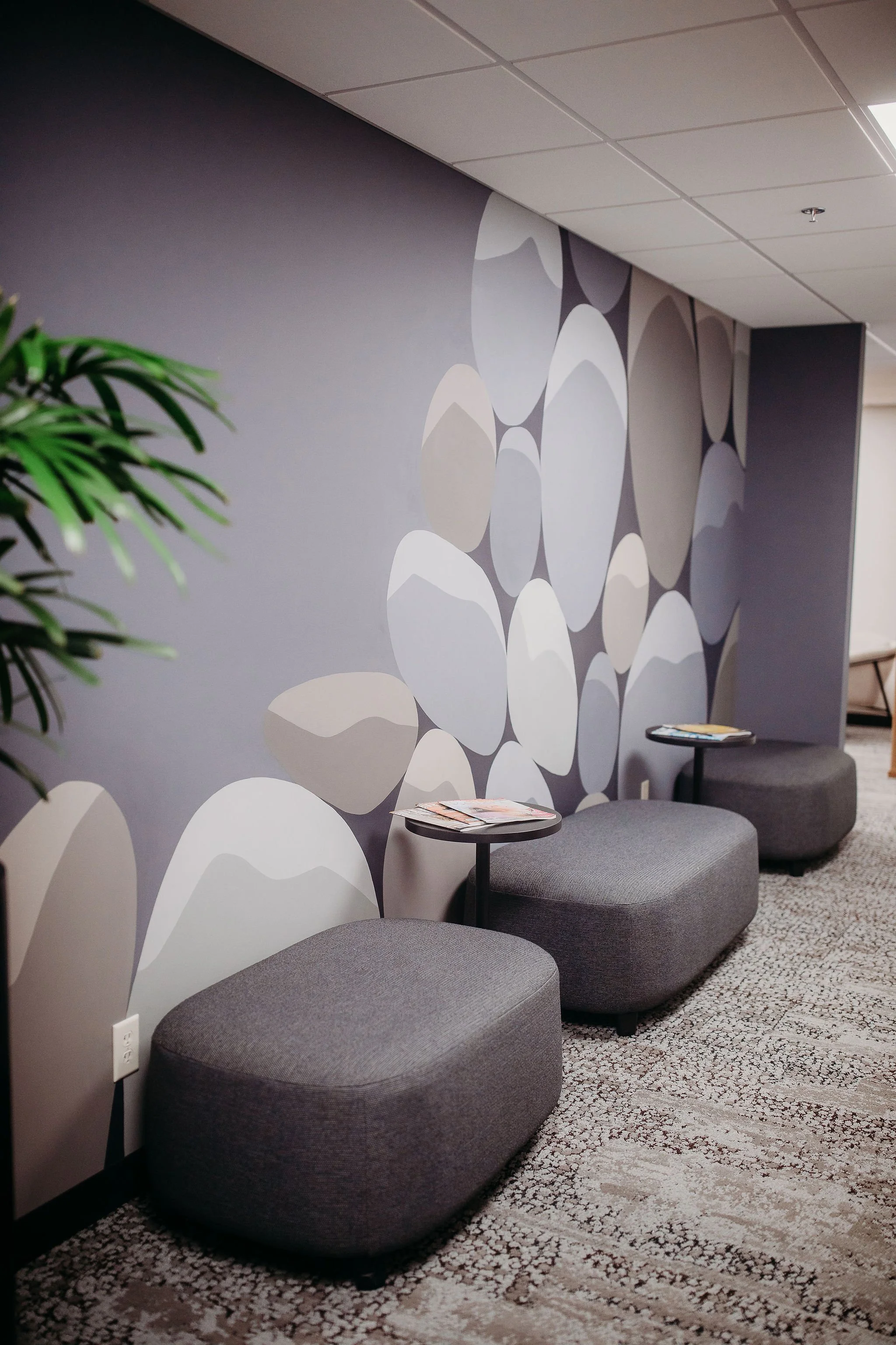 Contemporary lounge area with three gray upholstered seats, two small side tables with magazines, a wall with abstract art featuring large oval shapes in muted tones, part of a potted plant, and a patterned carpet.