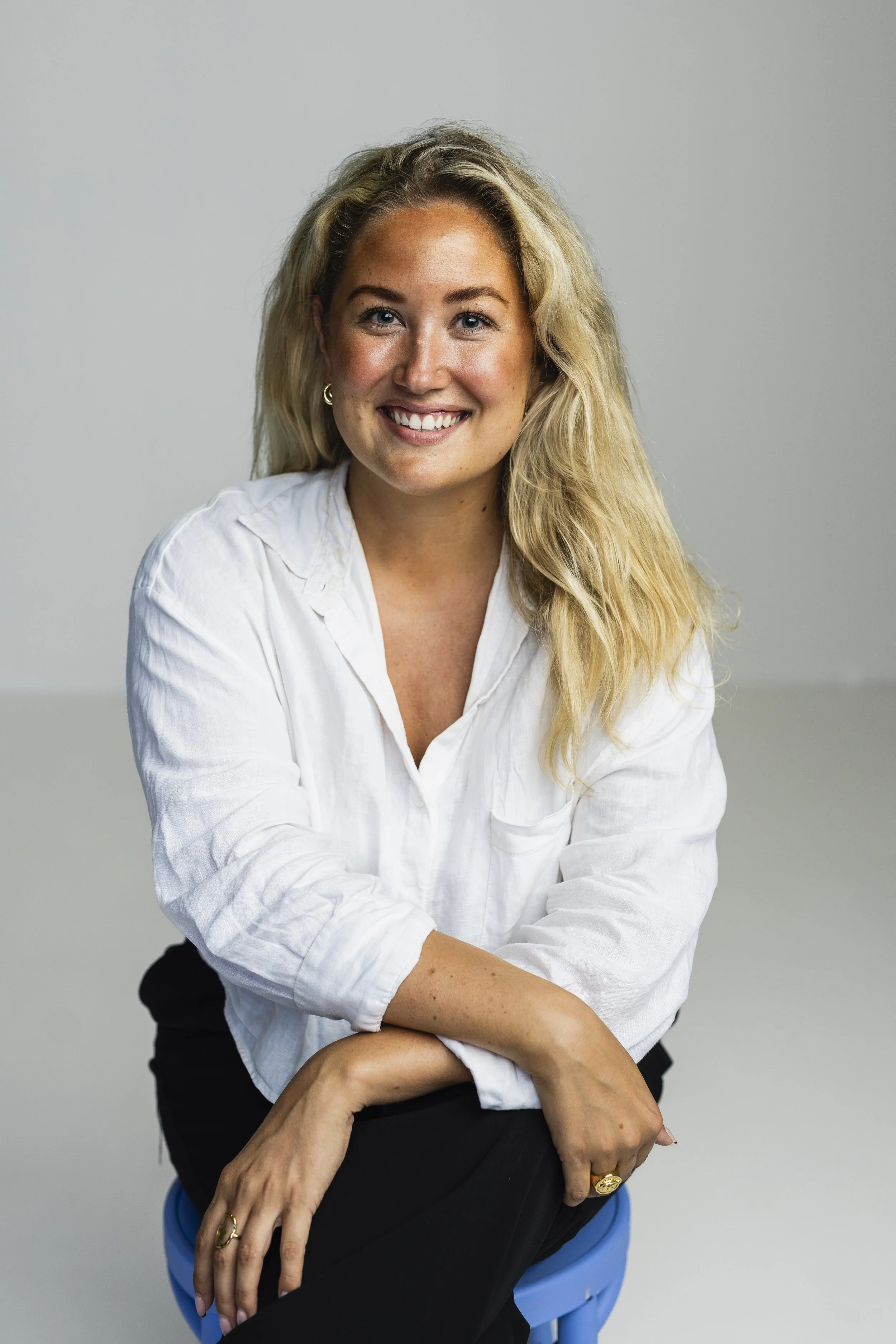 A smiling woman with blonde hair, wearing a white shirt, seated on a blue stool against a plain background.