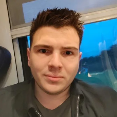 Portrait d'un jeune homme avec cheveux bruns, portant une veste noire, à l'intérieur près d'une fenêtre avec vue sur le ciel bleu