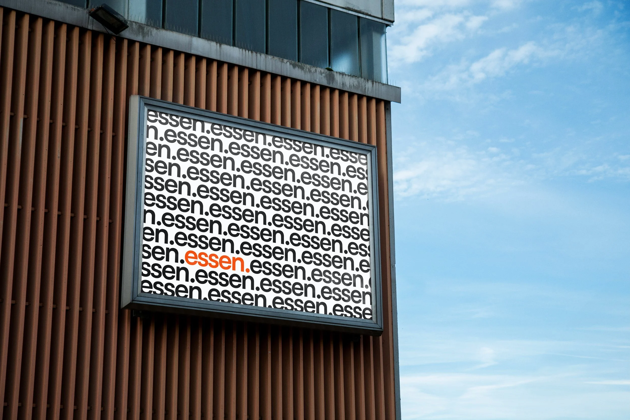 Affiche publicitaire avec le mot 'essen' répété en noir sur fond blanc, sauf un mot 'essen' en rouge, sur un mur en bois avec un ciel bleu en arrière-plan.