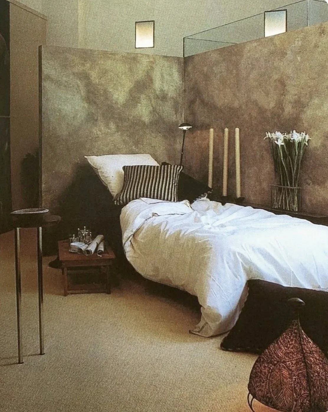 A cozy bedroom with a bed featuring white bedding and black and striped pillows, set against a textured beige wall with a modern black floor lamp, candles, and a vase of white flowers.
