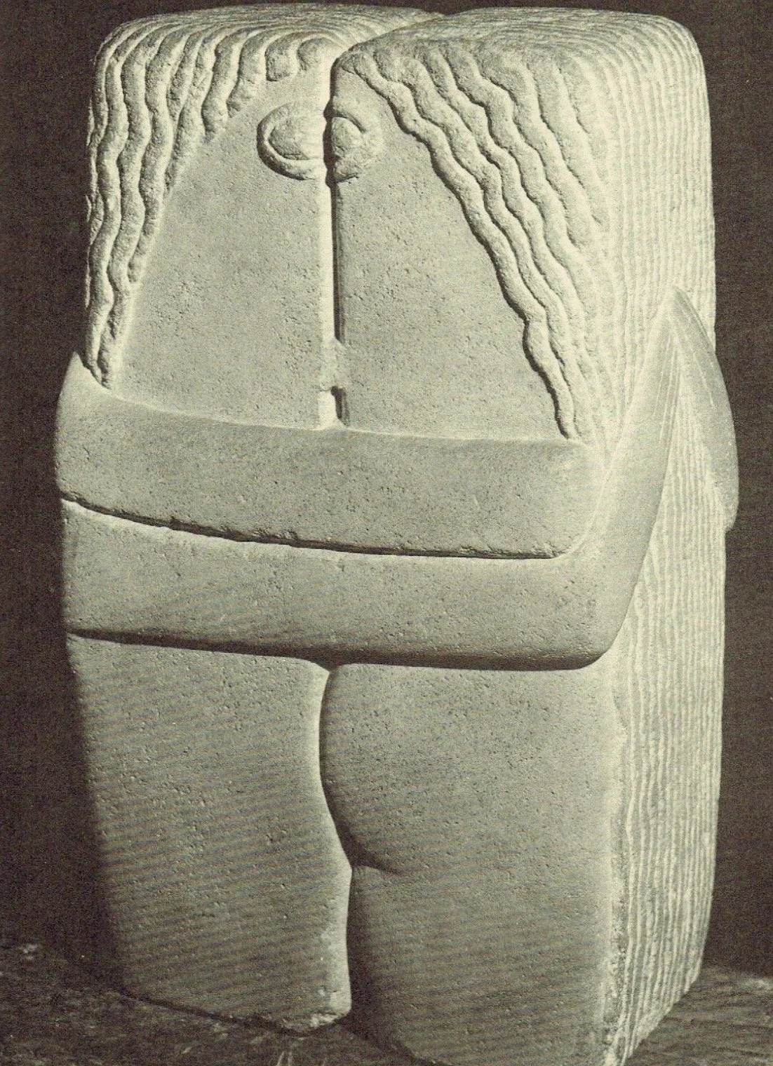 Ancient stone carving depicting two abstract human figures facing each other, with wavy hair and facial features, carved on a large rectangular stone block.