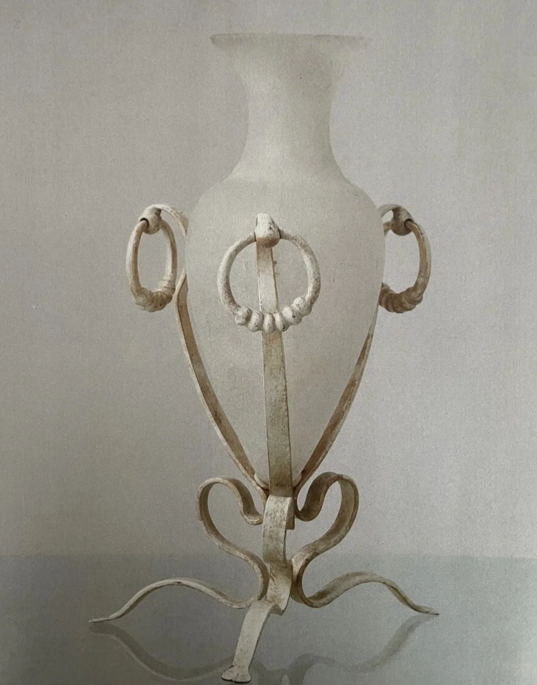 Vintage white metal vase with ornate handles and decorative legs, set against a plain background.