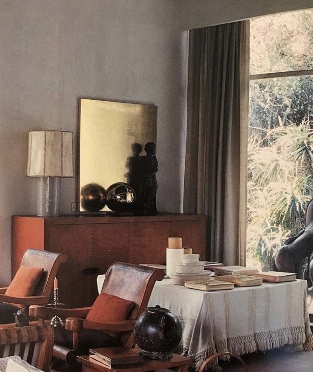 Interior of a living room with wooden armchairs, a side table, and a table with stacks of books and candles, by a large window with curtains showing outdoor foliage.
