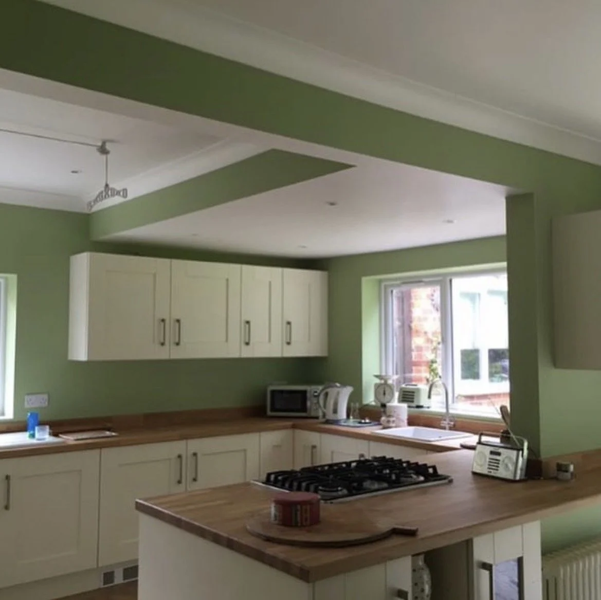 Kitchen with green walls, white cabinets, wooden countertops, a window, and appliances including a microwave, kettle, toaster, and gas stove.