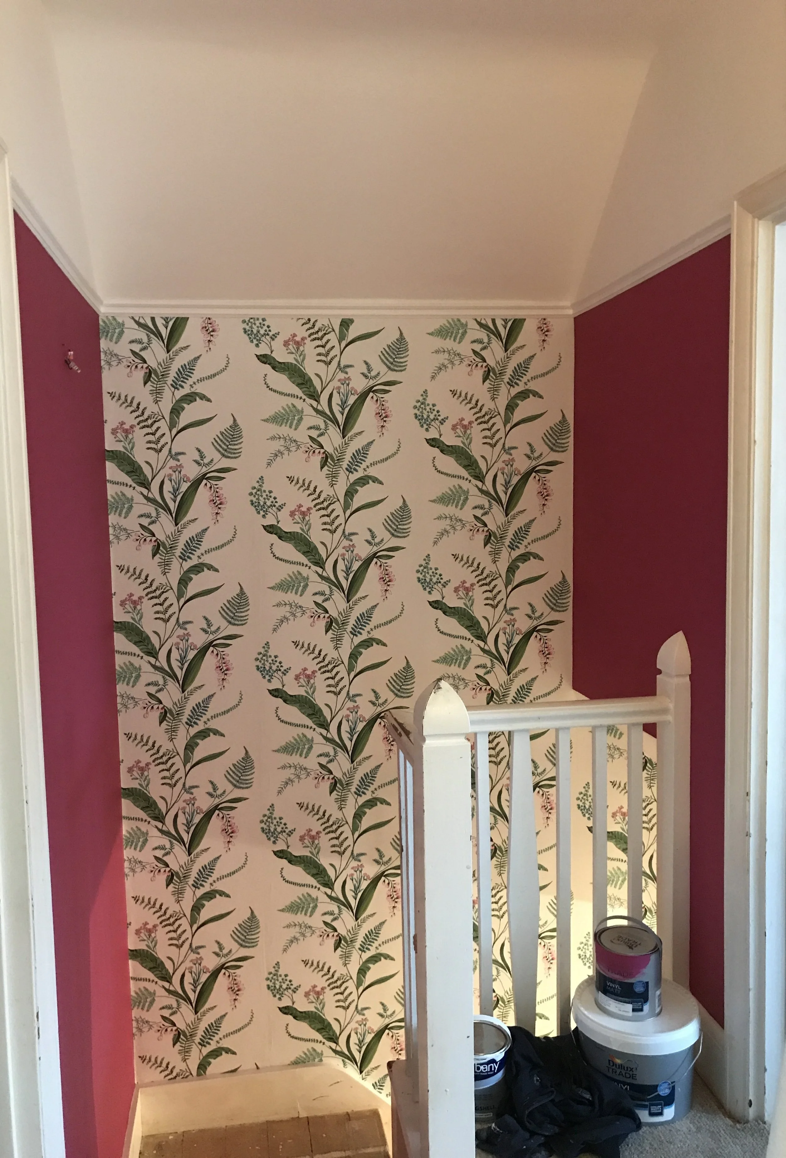 A small hallway corner with a floral wallpaper feature wall, maroon-colored side walls, and a white wooden railing. There are cans of paint, a cloth, and a container on the floor.