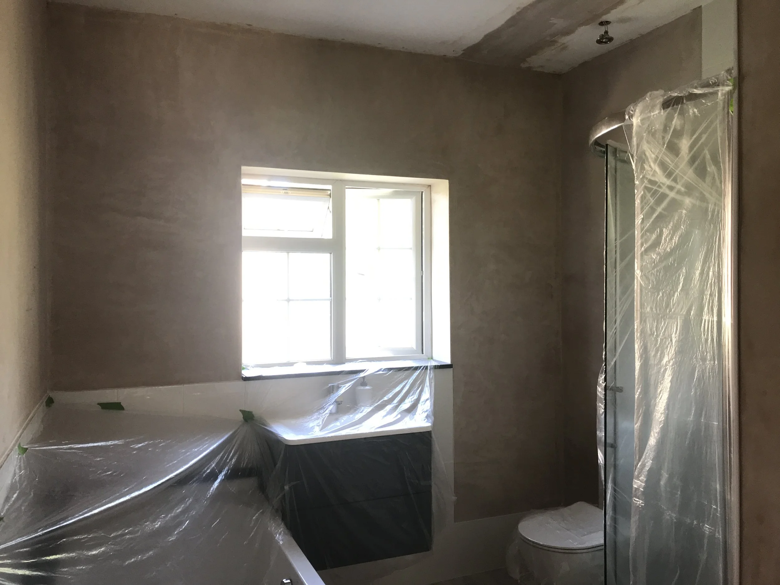 A bathroom under renovation with plastic coverings on the bathtub, shower, and toilet, and a window providing natural light.