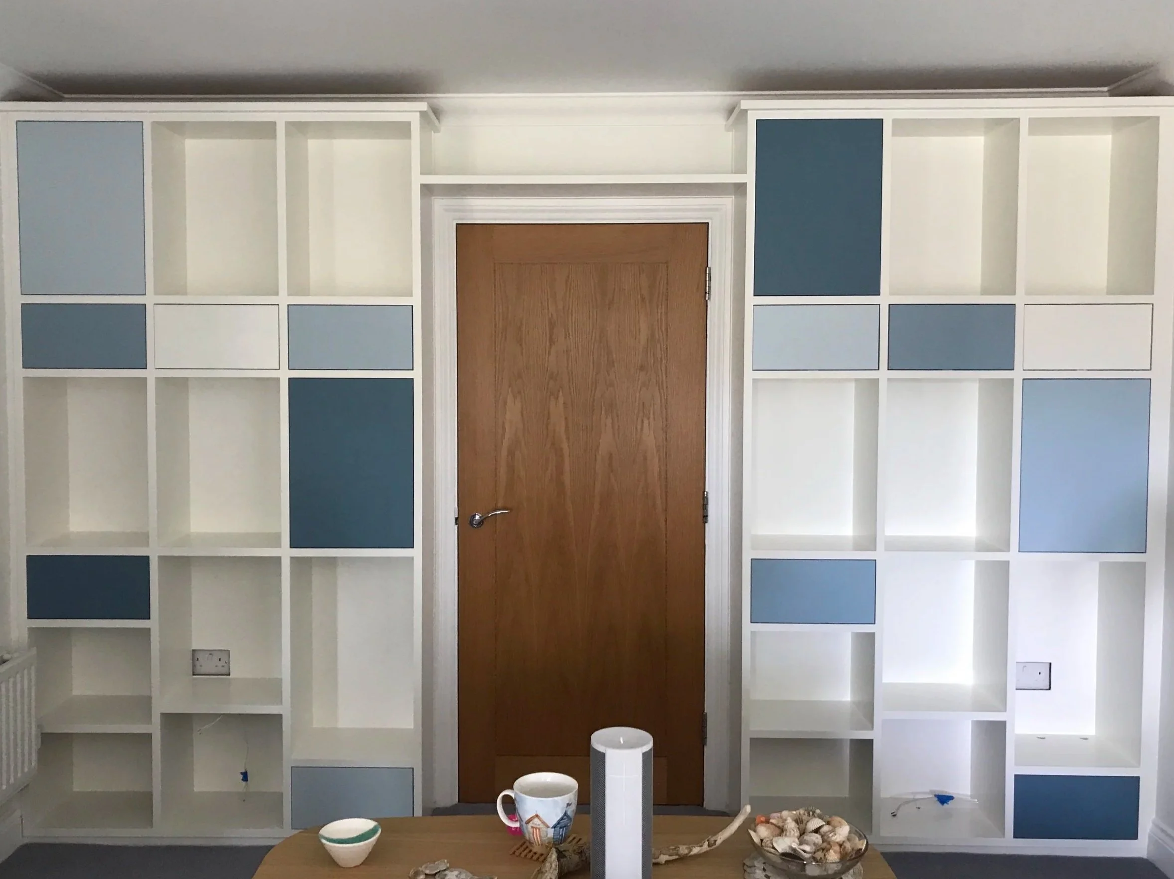 Empty white bookshelves with some blue accents surrounding a closed wooden door, with a table in front holding a mug, small bowl, and a bowl of shells.