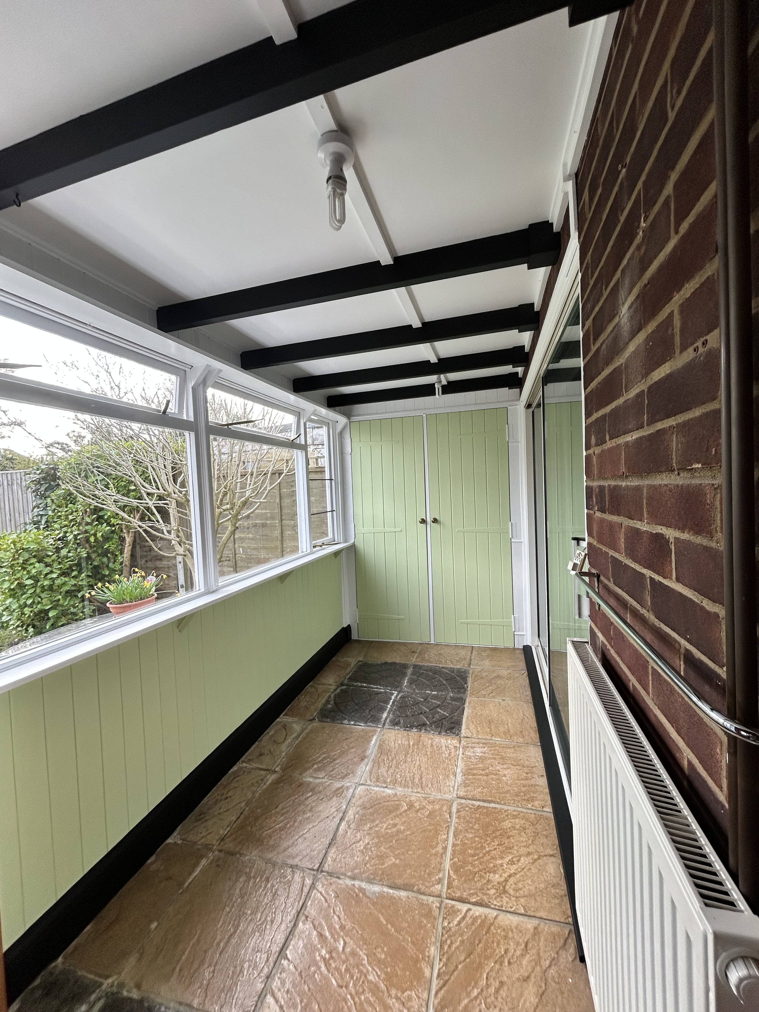 A small enclosed sunroom with large windows, green paneled walls, tiled floor, brick wall, radiator, and ceiling light fixture.