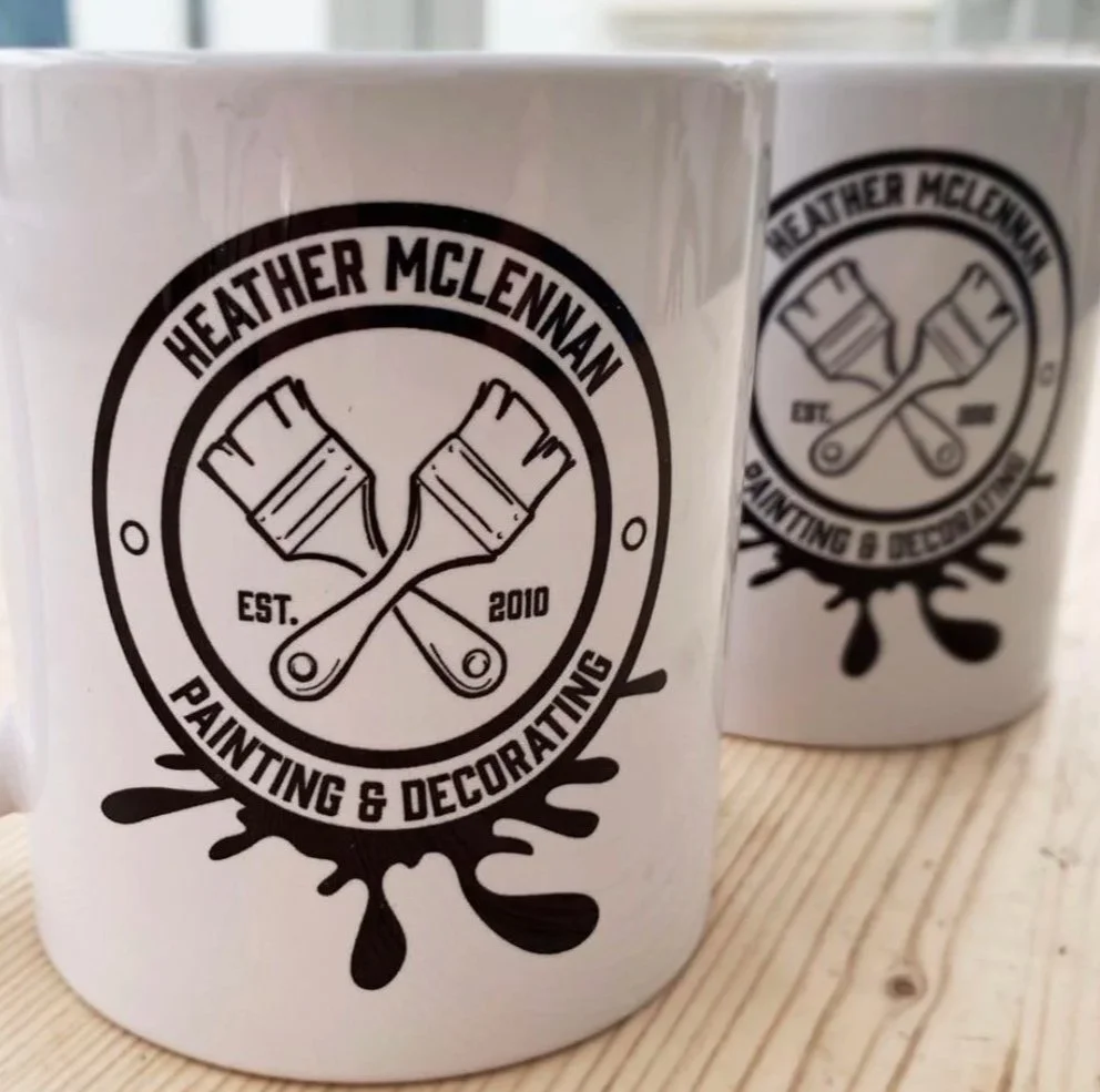 White mug with a black logo for Heather McLennan Painting & Decorating, featuring two crossed paintbrushes and text 'Est. 2010' on a wooden surface.