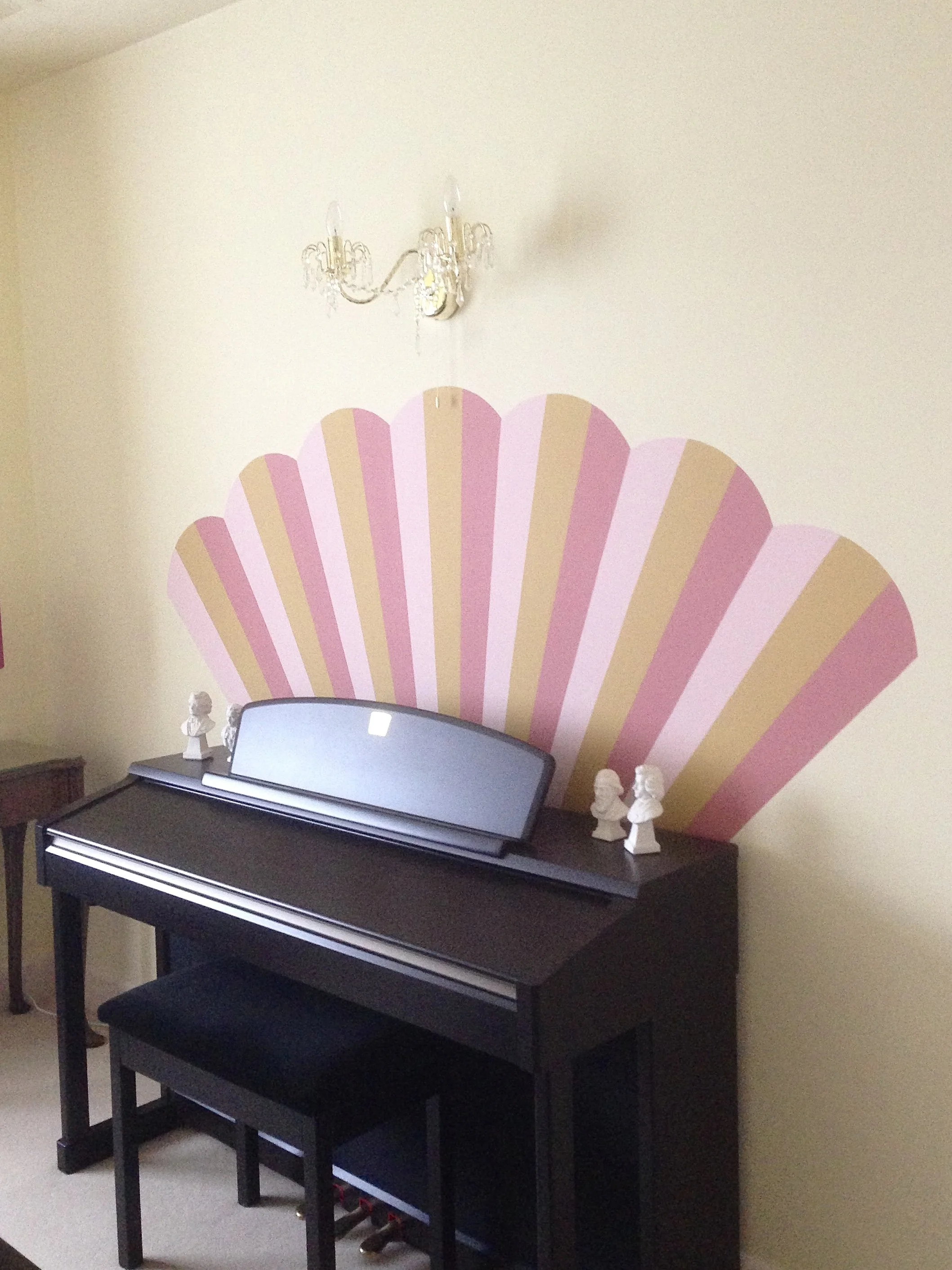 A black digital piano with a music stand and a matching black bench against a yellow wall, decorated with a pink and beige scalloped shell-shaped wall art. On top of the piano are four small white bust sculptures. There is a small wooden side table to the left of the piano and a crystal chandelier hanging from the ceiling.