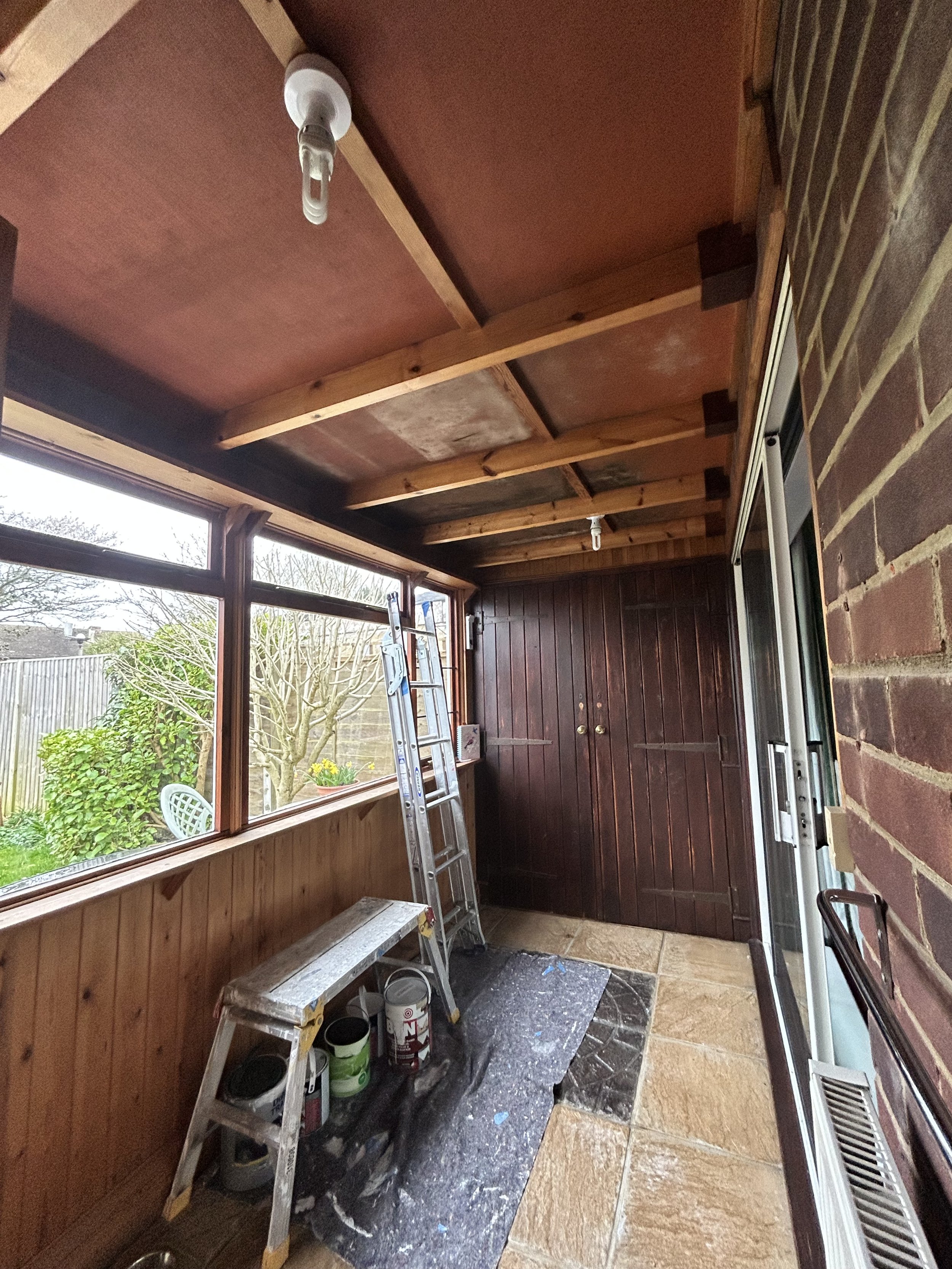 An enclosed porch or sunroom under renovation with a ladder, paint cans, supplies, and a wrapped bench, overlooking a garden outside