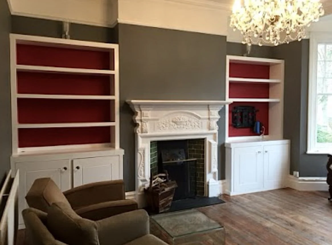 Living room with dark gray walls, white built-in shelves with red backs, a white ornate fireplace, a brown armchair, a small glass coffee table, and a large window. A chandelier hangs from the ceiling.
