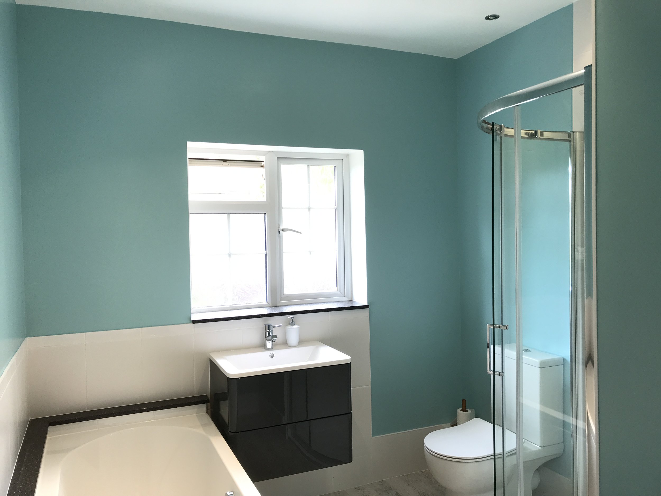 Modern bathroom with light blue walls, a window, a white and black vanity with a sink, a bathtub, and a partially visible toilet within a shower enclosure.