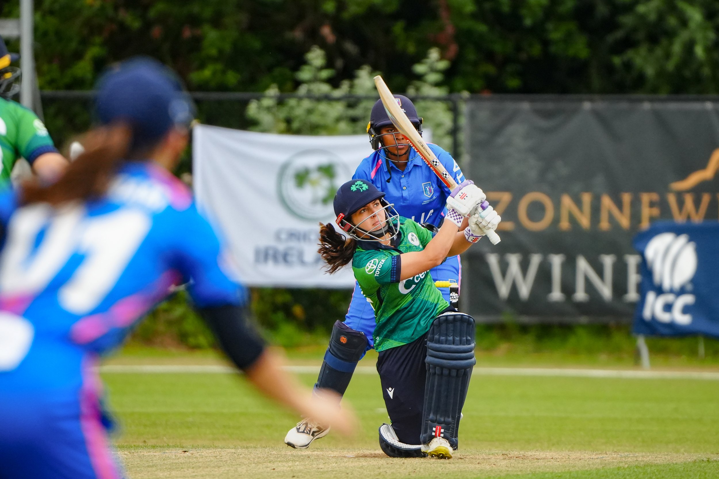 A female cricket player in a green jersey and black helmet is in the process of hitting the ball with her cricket bat during a match, with another female player in a blue jersey and helmet behind her, on a grass field with banners in the background.