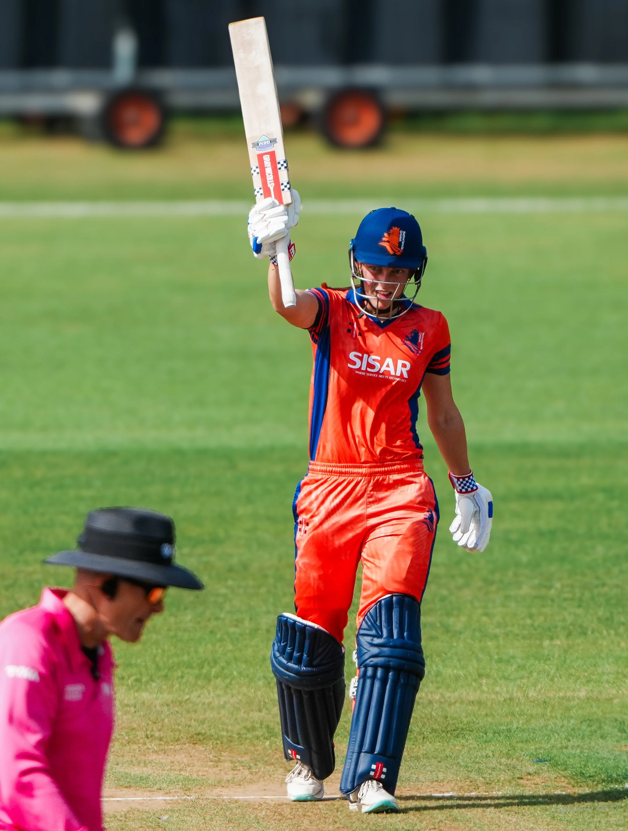 Cricket player in orange uniform celebrating with raised bat on the field, in front of a player in pink uniform and hat.