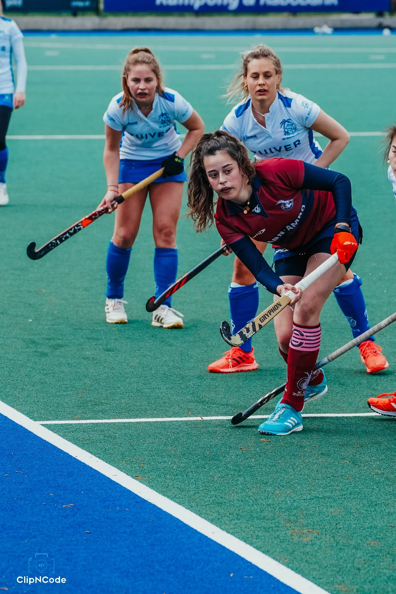 Women playing field hockey on a green turf, wearing sports uniforms with blue and maroon colors, and holding hockey sticks.