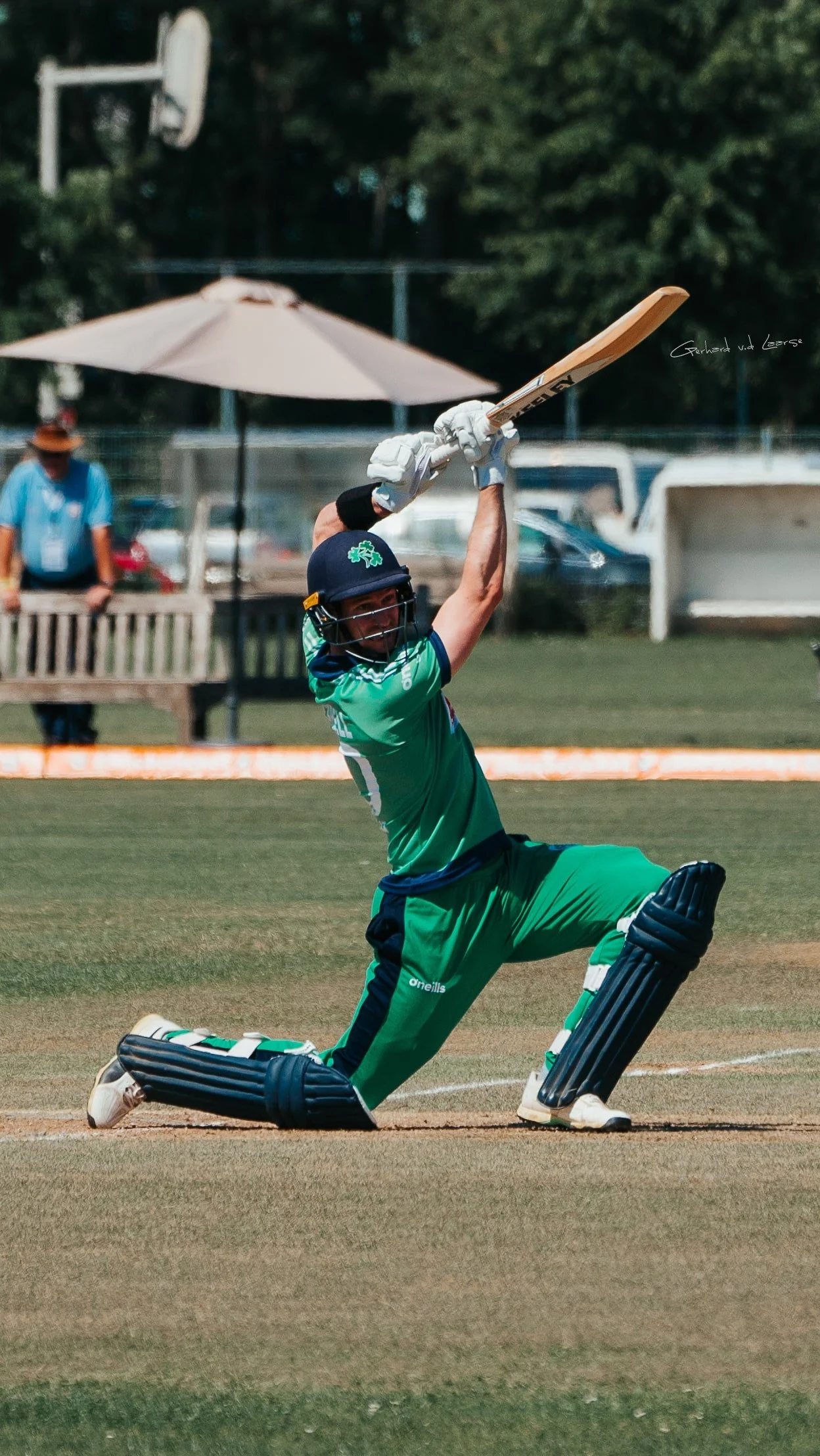 Cricket player in green uniform kneeling and hitting a ball with a bat on a cricket field.