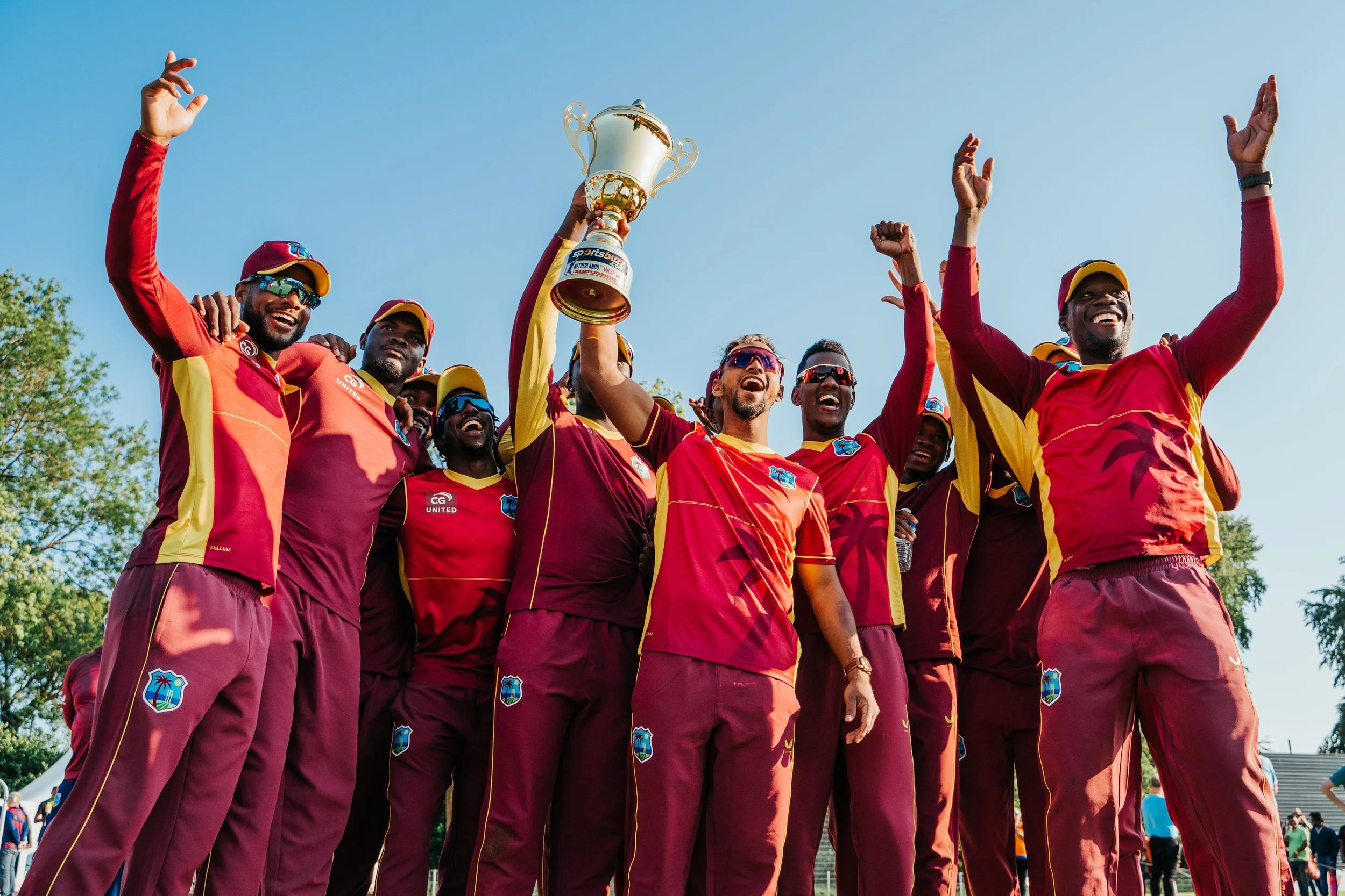 Cricket team in maroon and yellow uniforms celebrating victory, holding a trophy, outdoors on a sunny day.