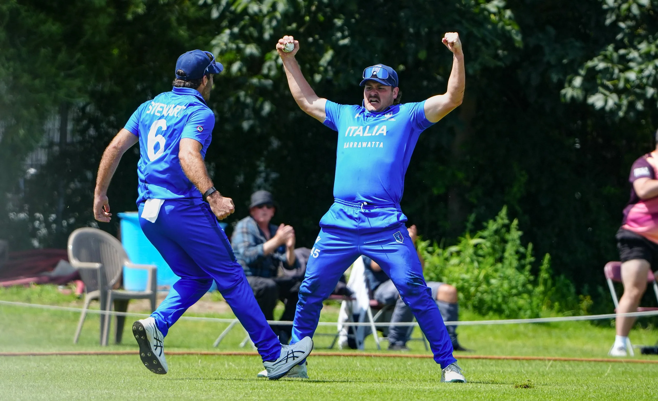 Two cricket players in blue uniforms celebrating on the field. One with his arms raised, the other with a clenched fist. Spectators sit in the background.