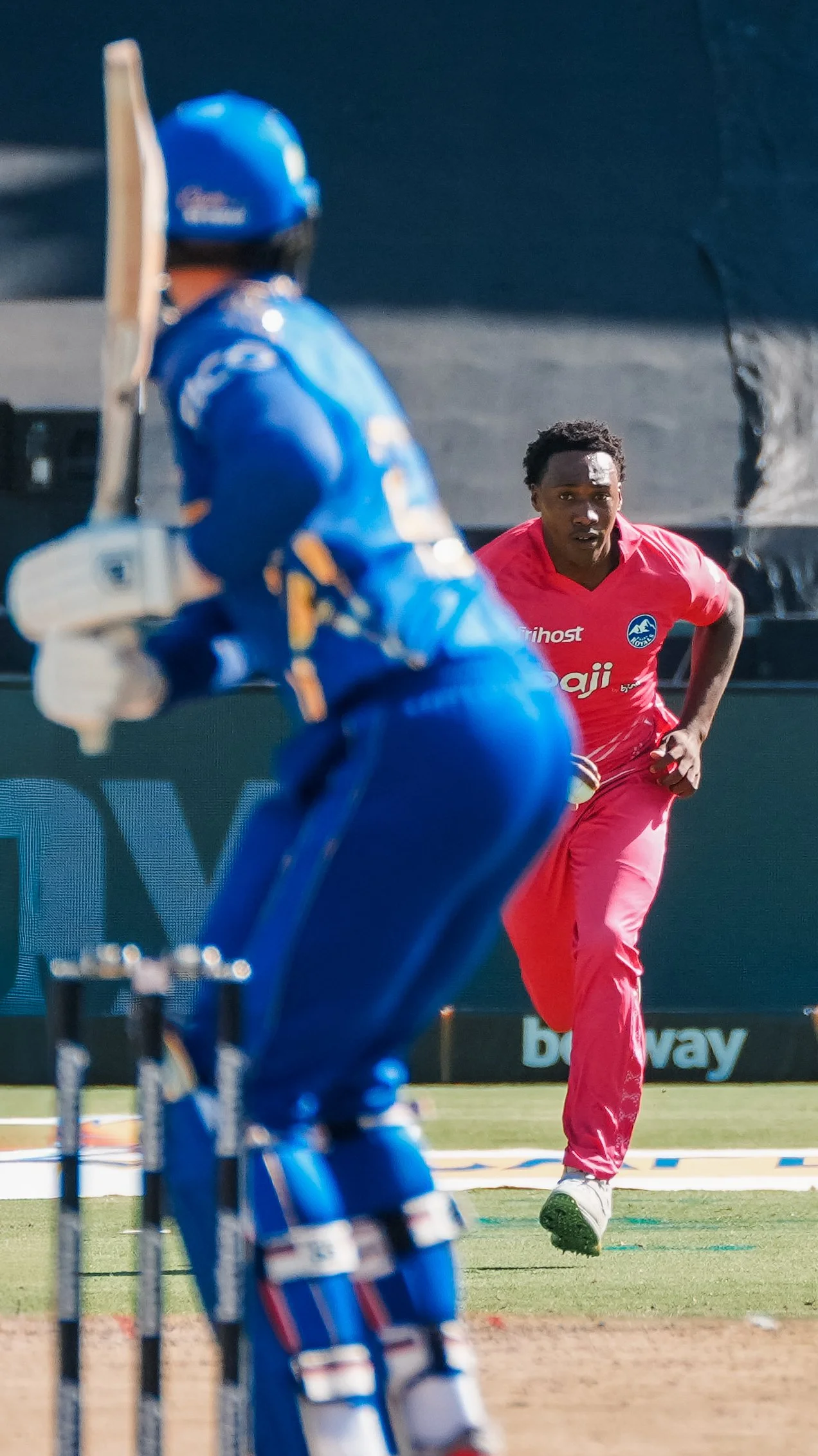 A cricket player in a red uniform running towards the pitch while a wicketkeeper in a blue uniform and protective gear stands behind the stumps on the cricket field.