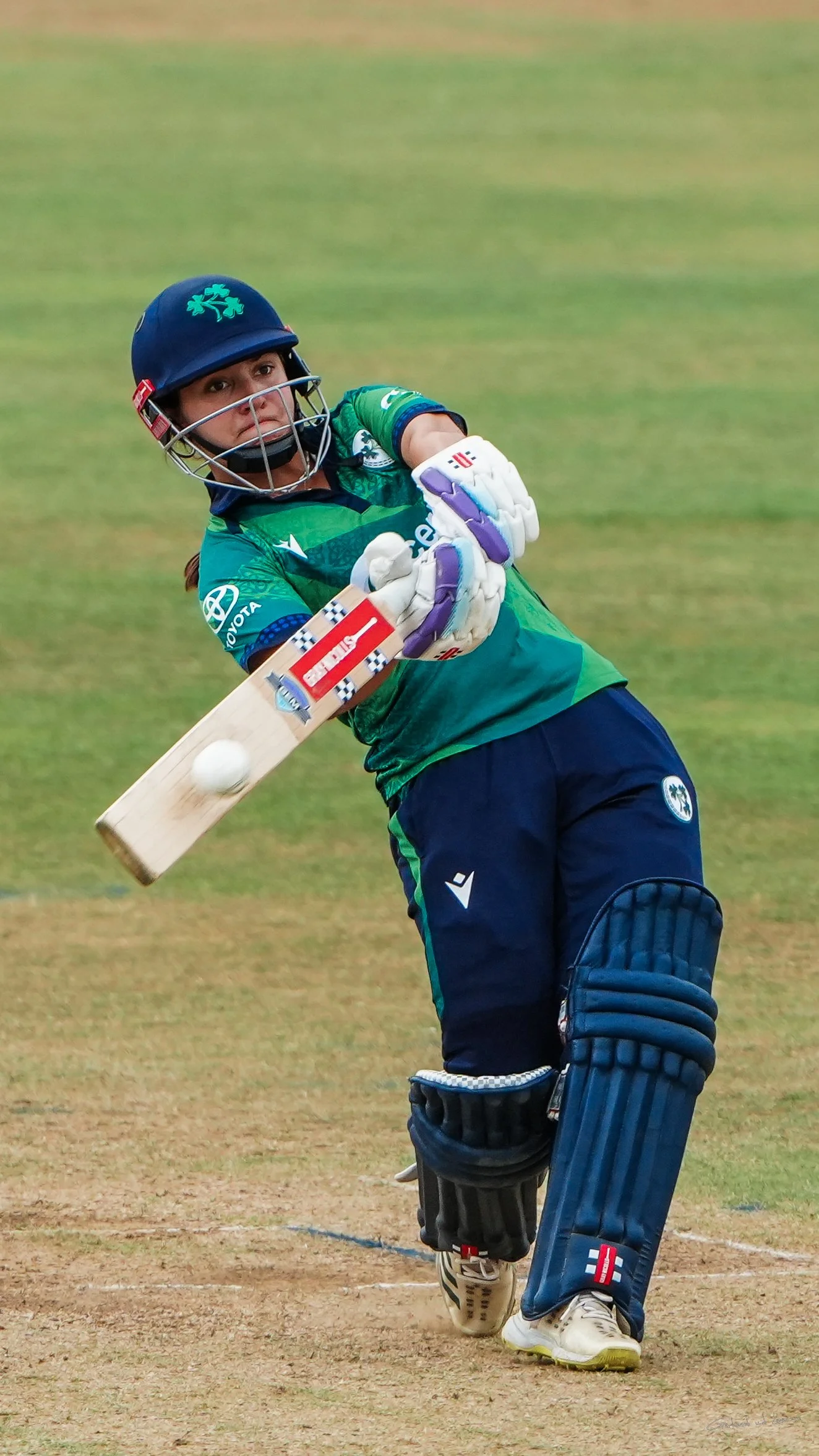 A woman cricketer batting on a cricket field with green grass.
