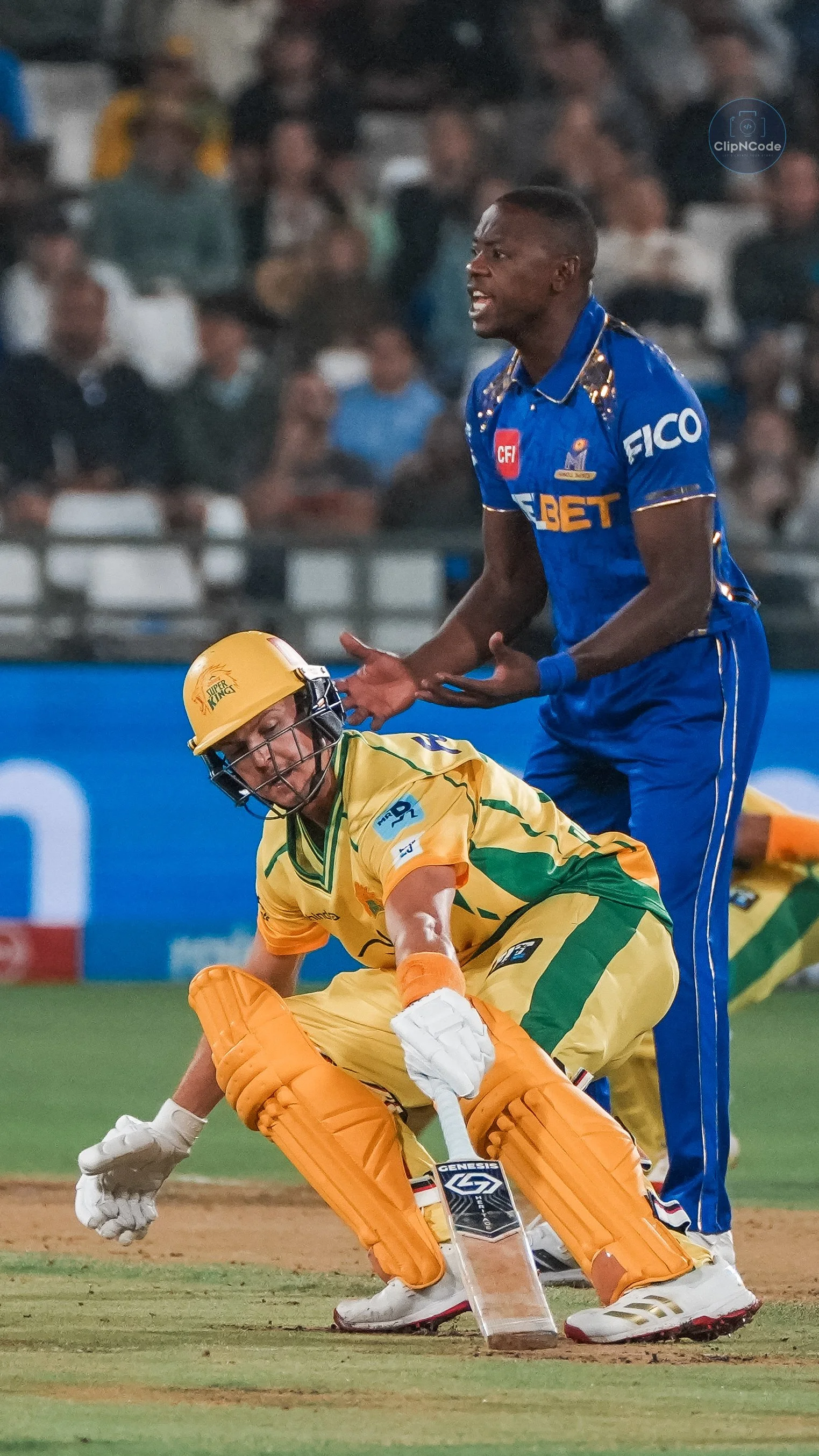 A cricket player in yellow protective gear and a helmet is on the ground, while an opposing player in a blue jersey gestures in frustration during a match with a crowd in the background.