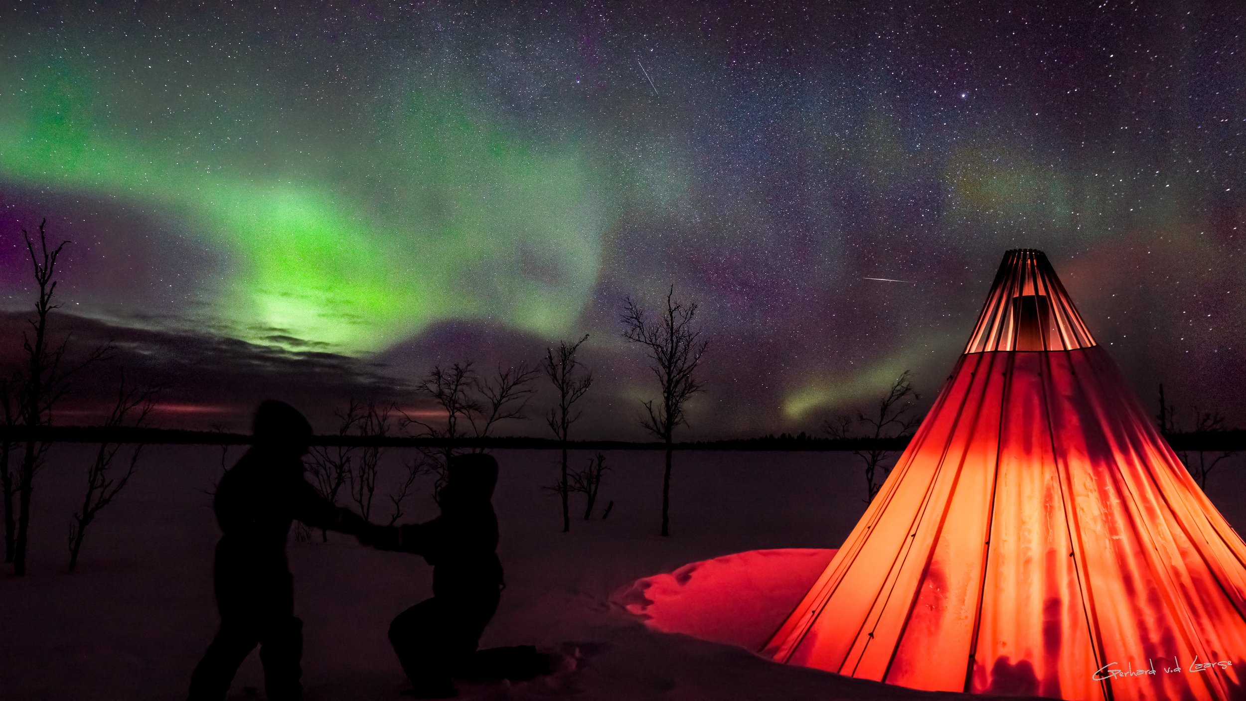 A night scene with the Northern Lights in the sky, a starry sky, a glowing red tipi, and silhouetted people in a snow-covered landscape with leafless trees.