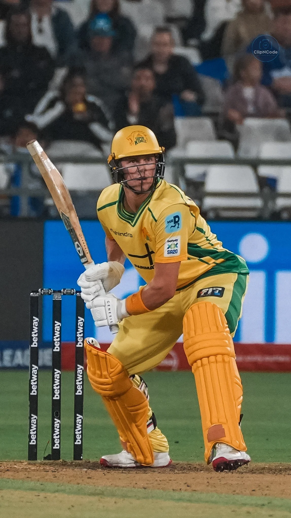 A cricket player in yellow and green uniform is behind the stumps, holding a bat, ready to defend or hit the ball during a match.