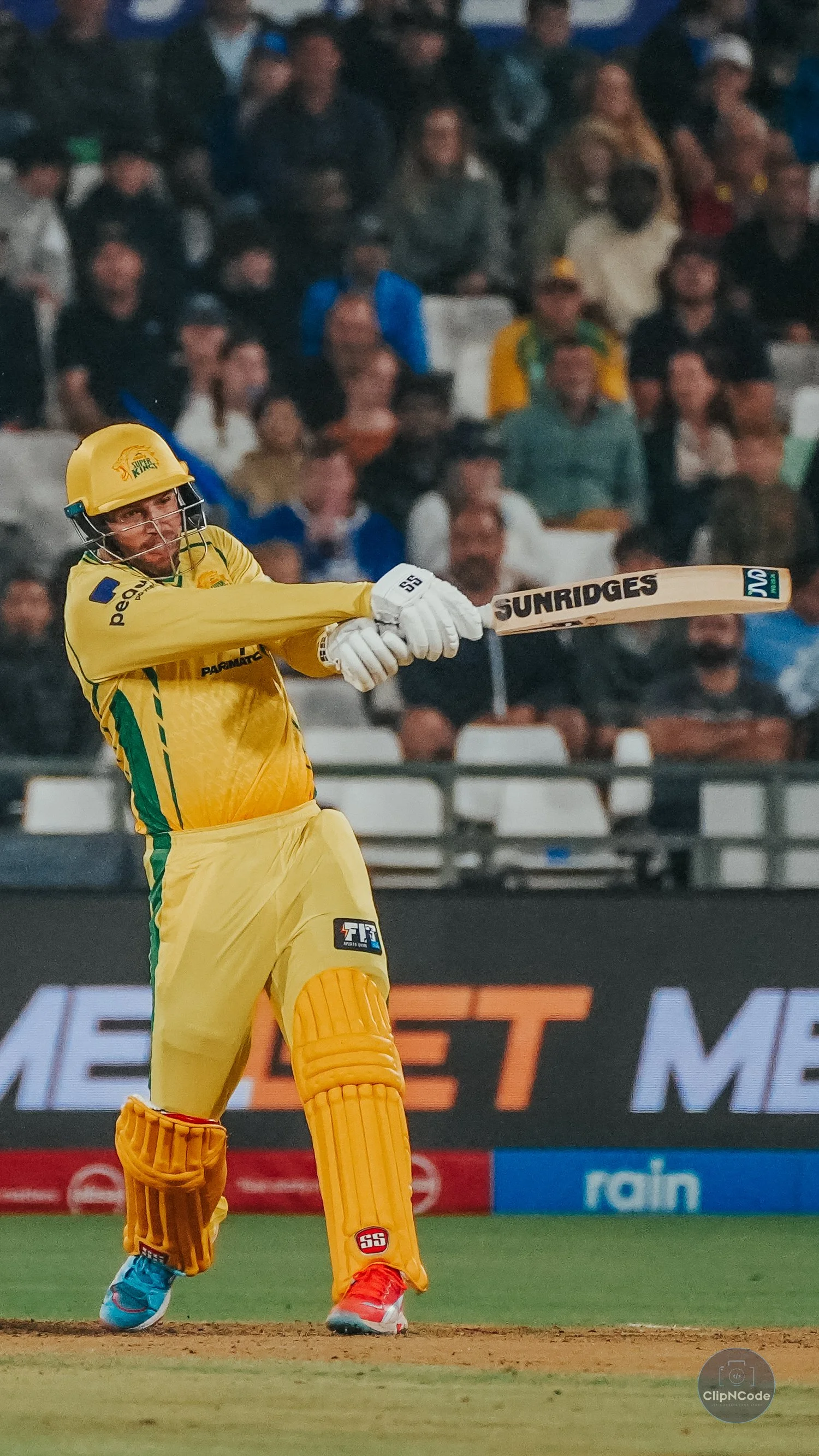Cricket player in yellow uniform swinging a bat with an audience in the background.
