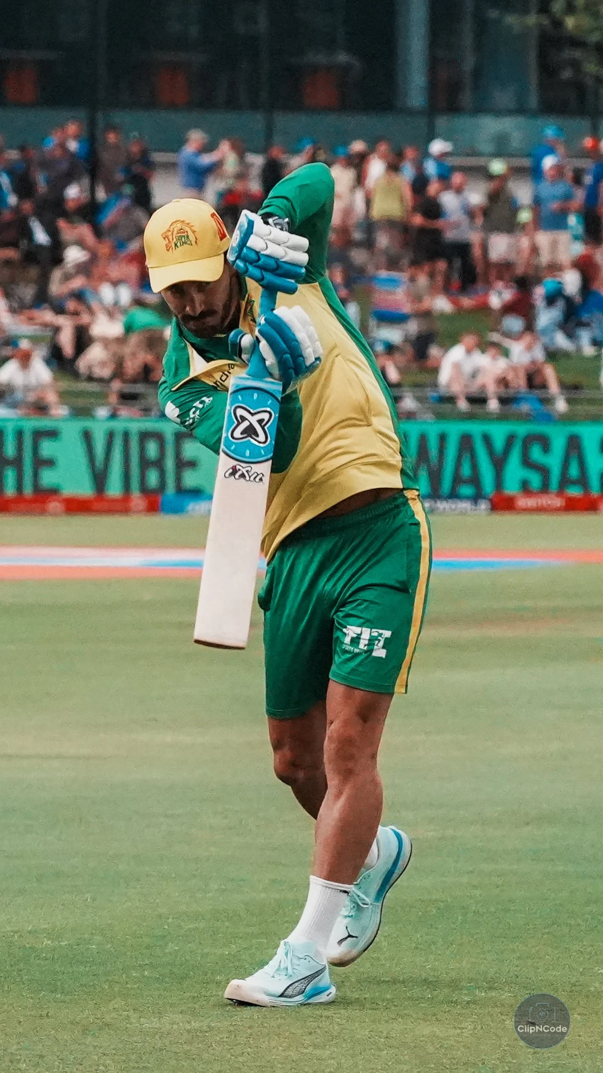 Cricket player in green and yellow uniform, wearing a yellow cap, in action on the field during a match with spectators in the background.