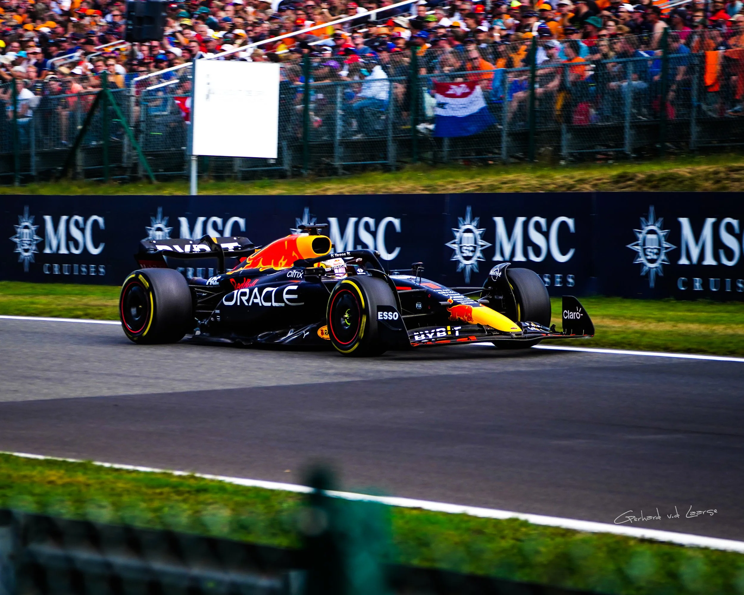 A black Formula 1 race car with Red Bull branding on a race track, with a crowd of spectators behind a fence in the background.