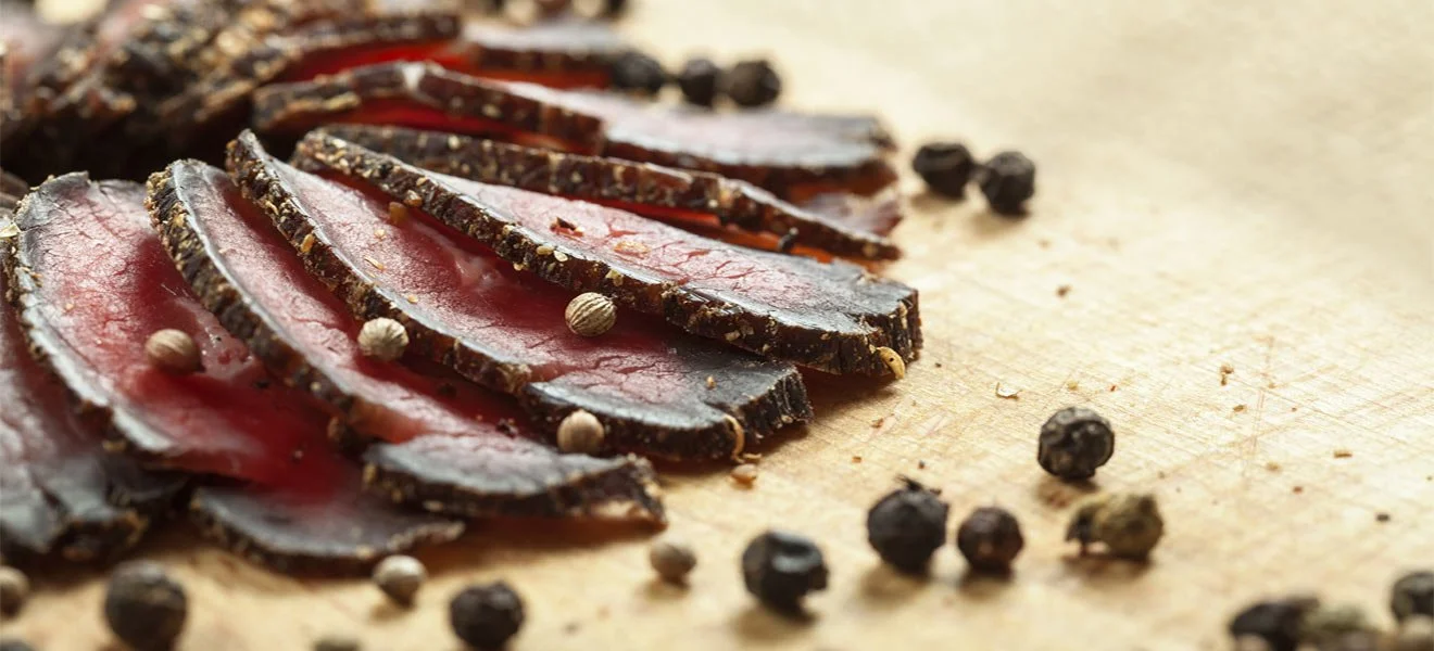Close-up of sliced cured meat with black peppercorns on a wooden cutting board.