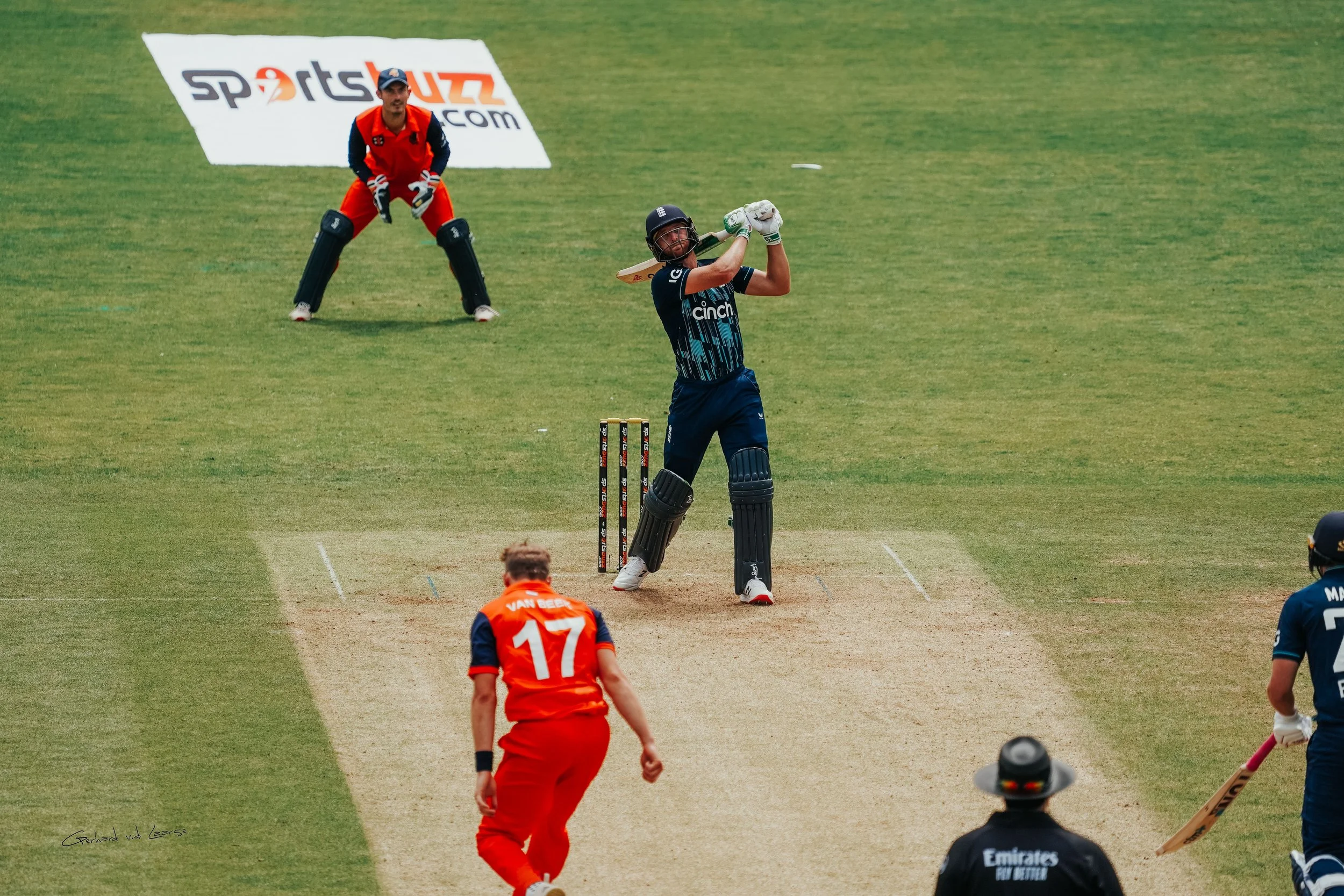 A cricket player in navy blue uniform is batting, with the ball in mid-air near his bat. An orange team player is running towards him. An umpire in black attire and a hat is standing in front of the pitch. The wicketkeeper and a fielder are behind, w