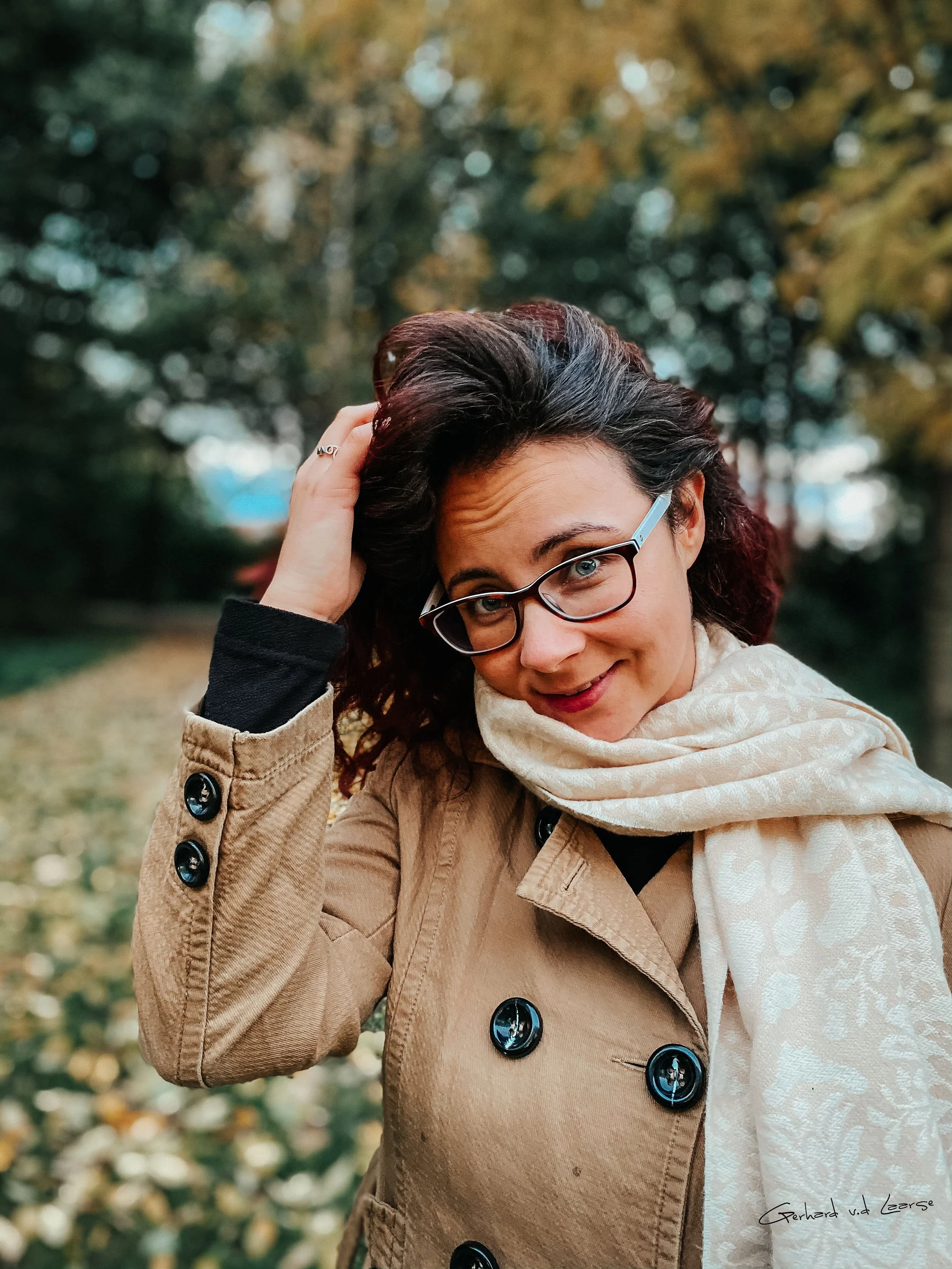 A woman with dark hair, glasses, and a beige coat, smiling outdoors during autumn, with trees and fallen leaves in the background.