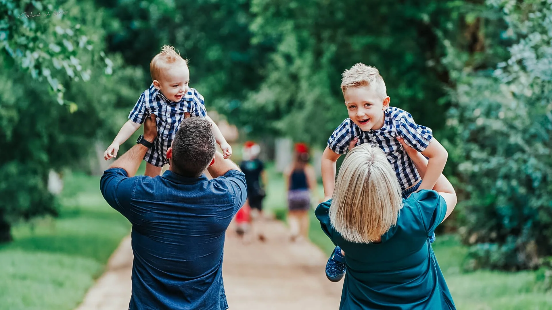 Two children, a boy and a girl, are being lifted into the air by their parents in a park with green trees. The children are smiling and wearing plaid shirts, while the parents are holding them up from behind.