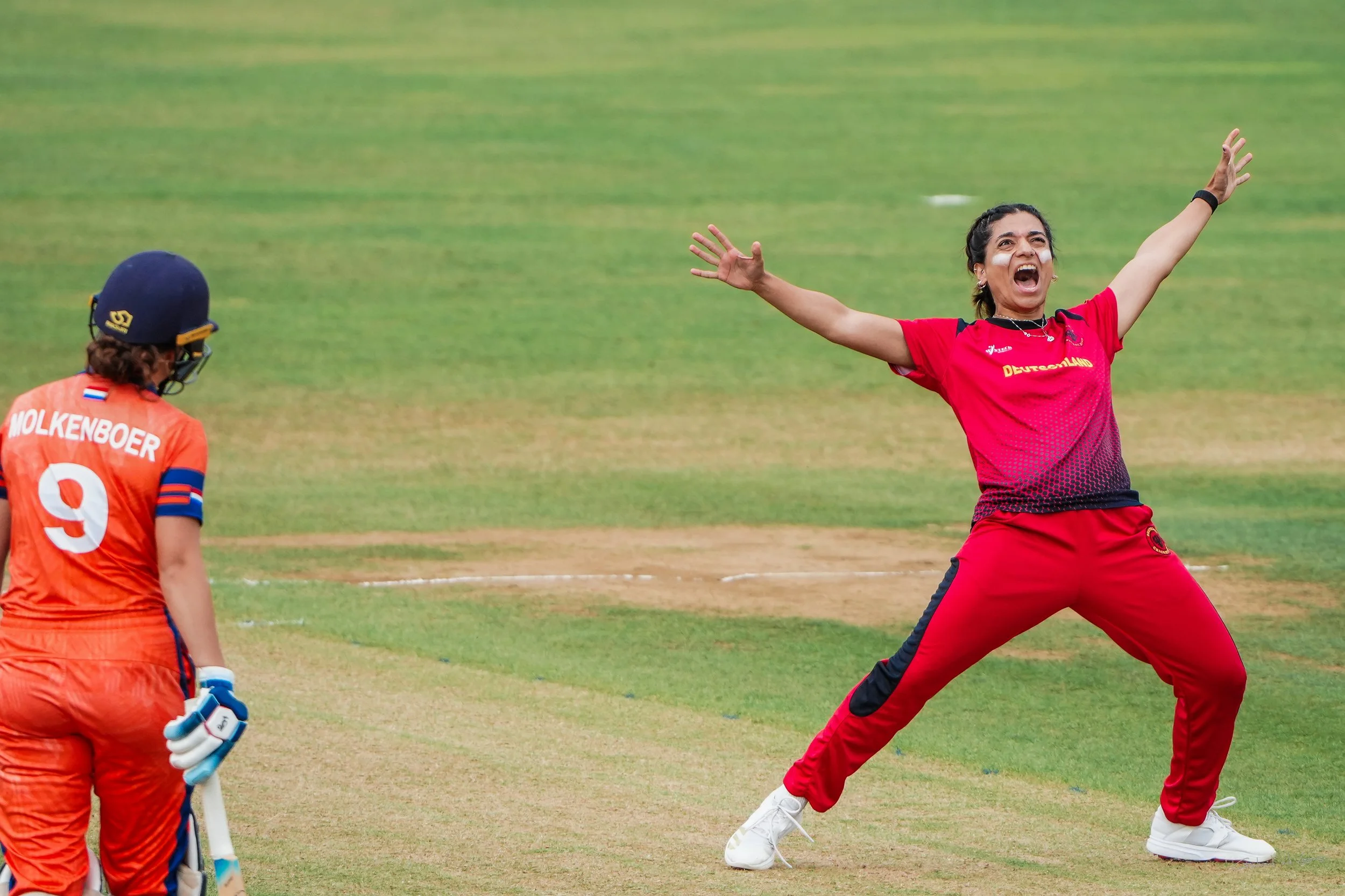 A female cricket player in red athletic wear with arms outstretched and mouth open, celebrating on the cricket field. Another female cricket player in orange uniform and helmet looks on.