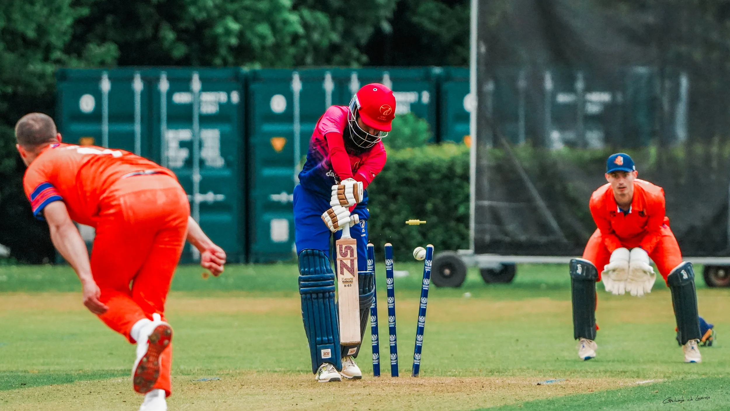 A cricket game in action, with a batsman in a red and blue uniform and red helmet preparing to hit the ball, two fielders in bright orange uniforms and helmets, and a cricket pitch with wickets and stumps on a grassy field.