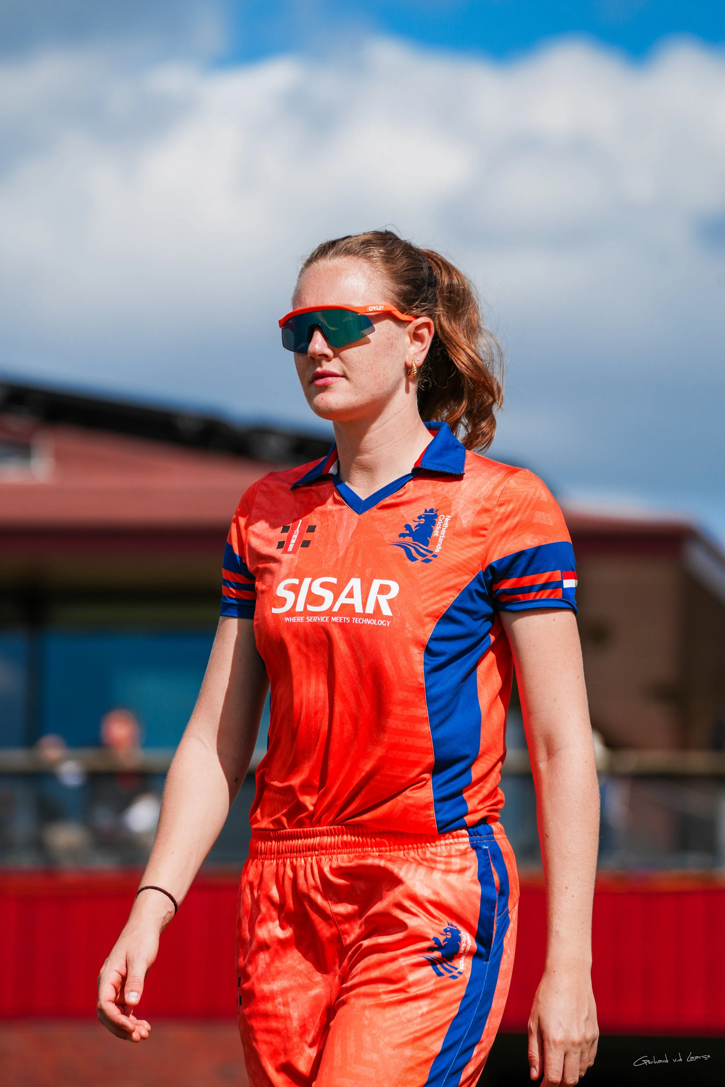 A female athlete wearing sunglasses and a red and blue sports uniform with the logo of the Dutch national team and sponsor text, walking outdoors under cloudy skies.