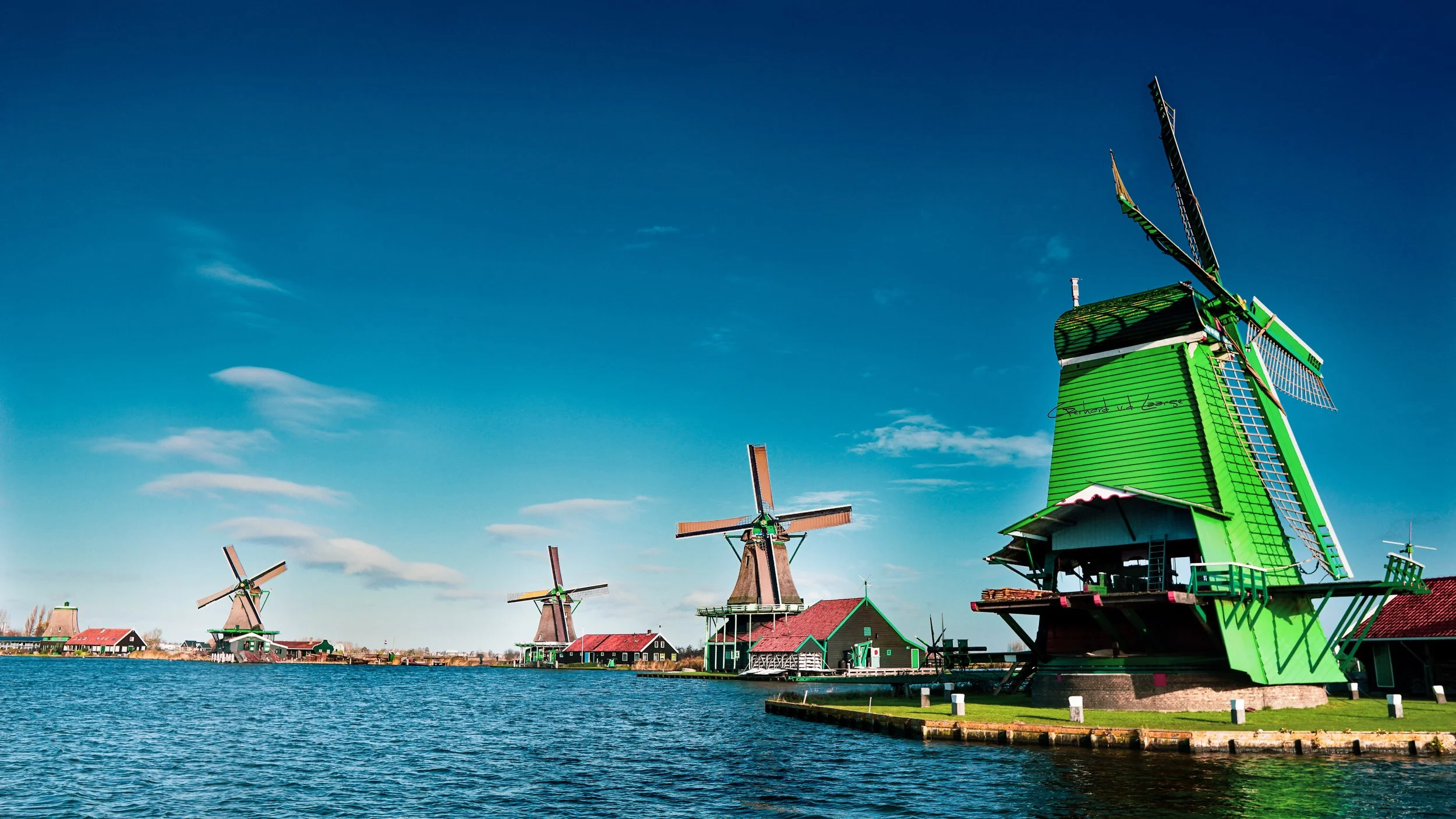 Multiple traditional Dutch windmills along a river under a bright blue sky with few clouds.