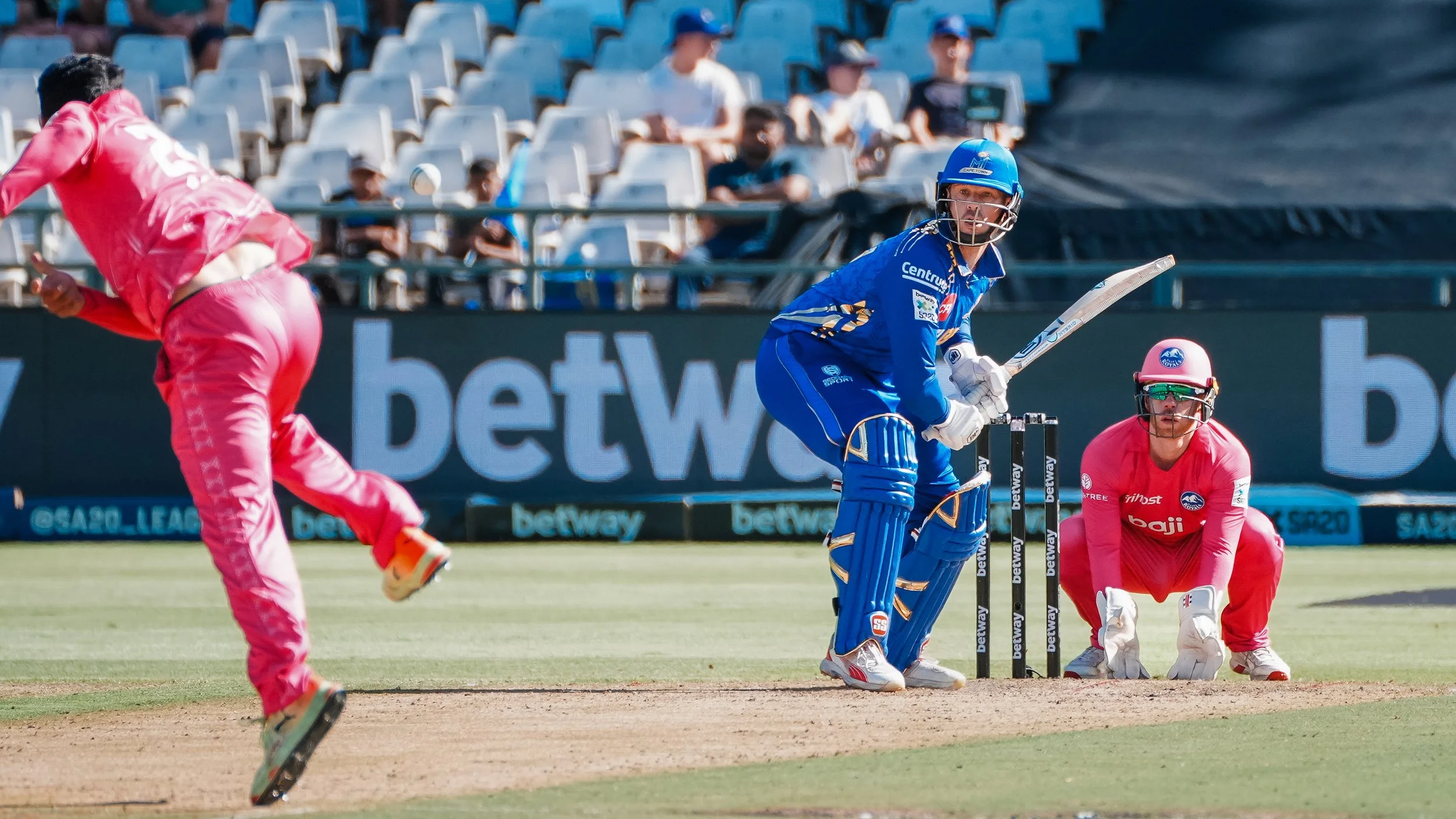 Cricket players in action during a match, with one player in blue preparing to hit the ball while a wicketkeeper in pink crouches behind the stumps and another player in pink runs toward the pitch.