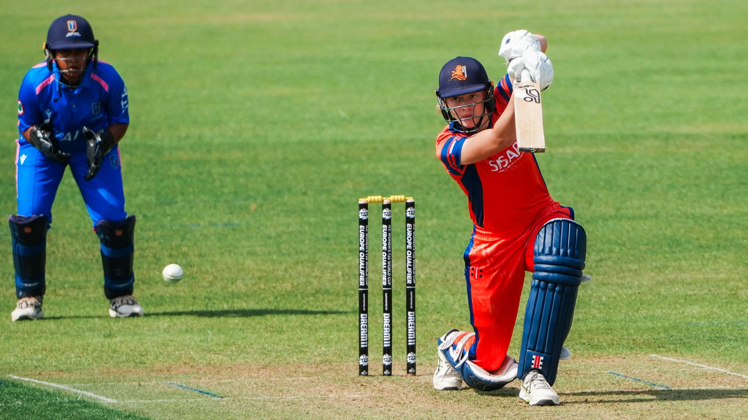 A female cricket player in a red uniform batting during a match, with a wicketkeeper in a blue uniform behind her and wickets on the pitch.