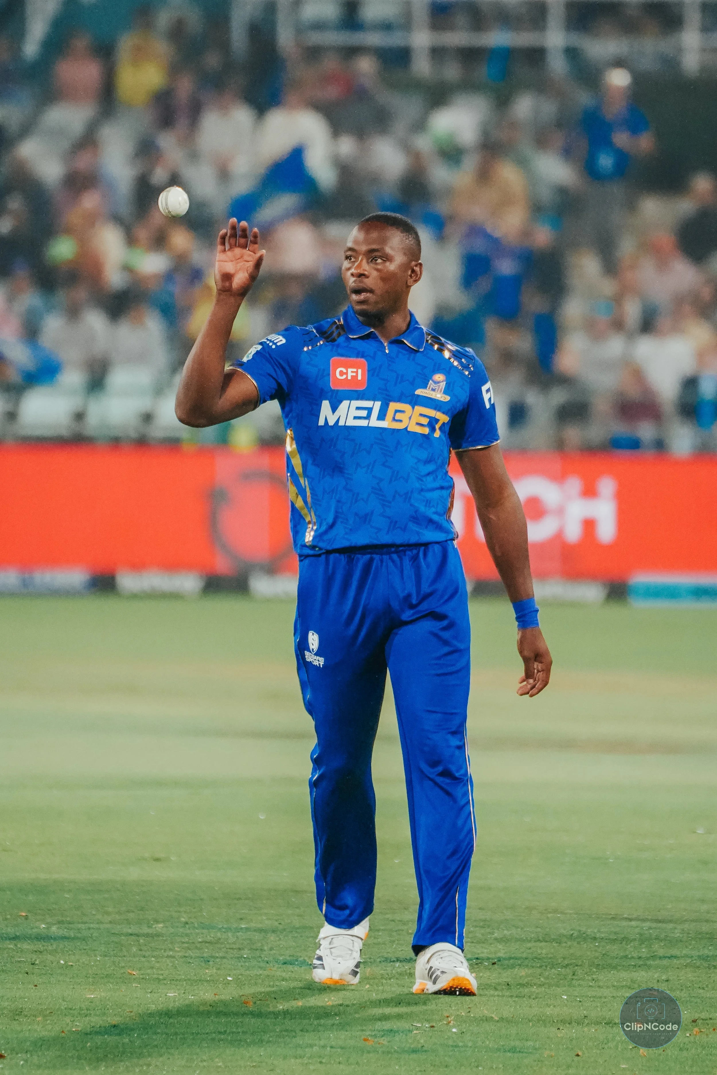 A cricket player in a blue uniform on a field, raising his right hand, with a crowd in the background.