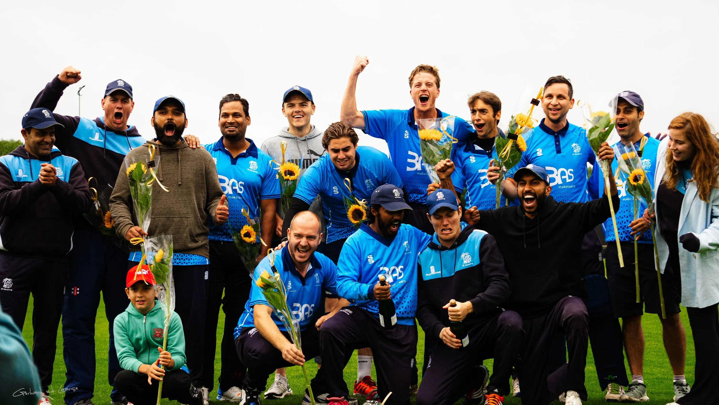 Cricket team celebrating victory, holding flowers and bottles on a grassy field.