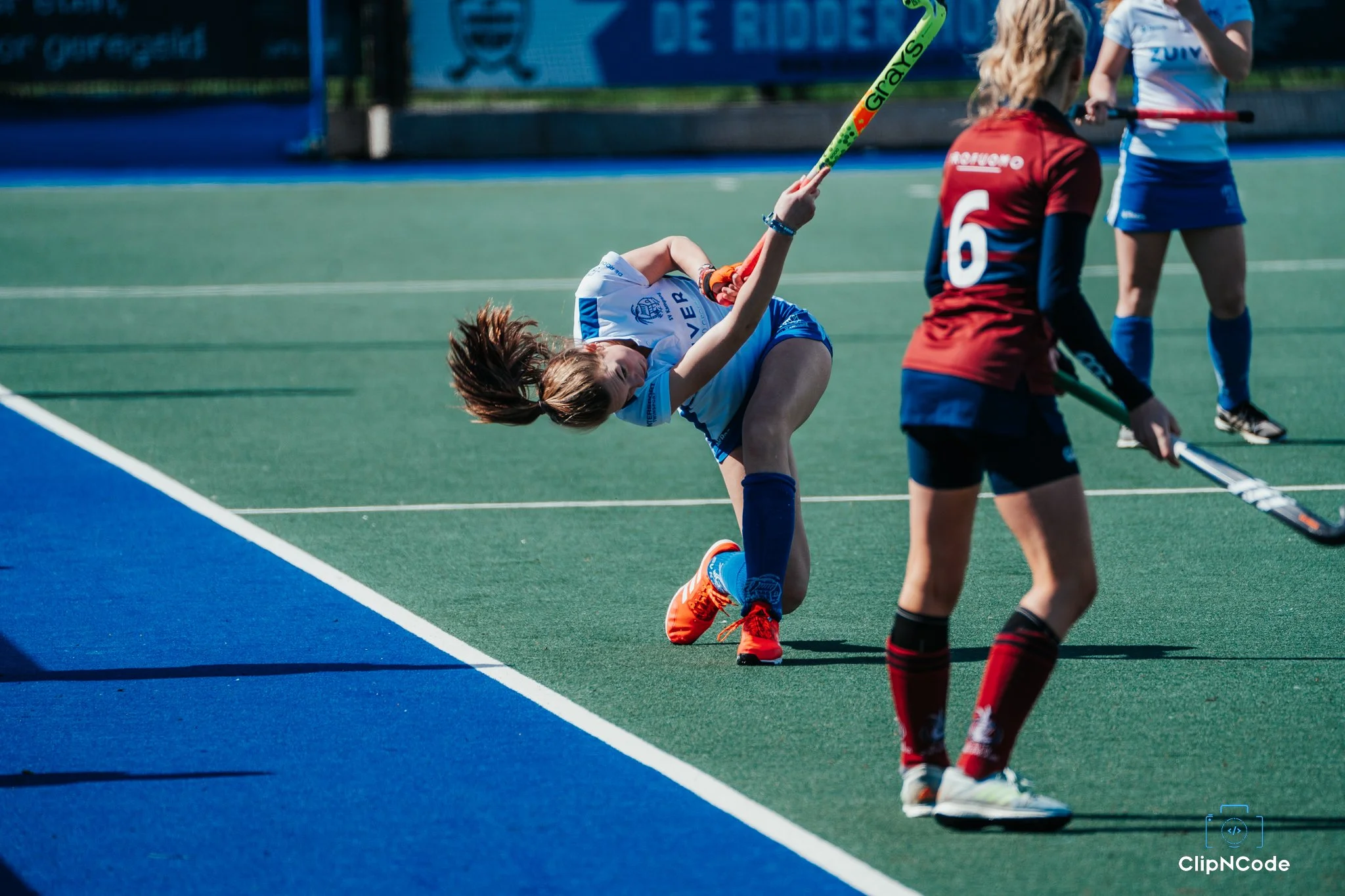 Female field hockey player in blue and white uniform falling to the ground during a game, with other players nearby on the green field.